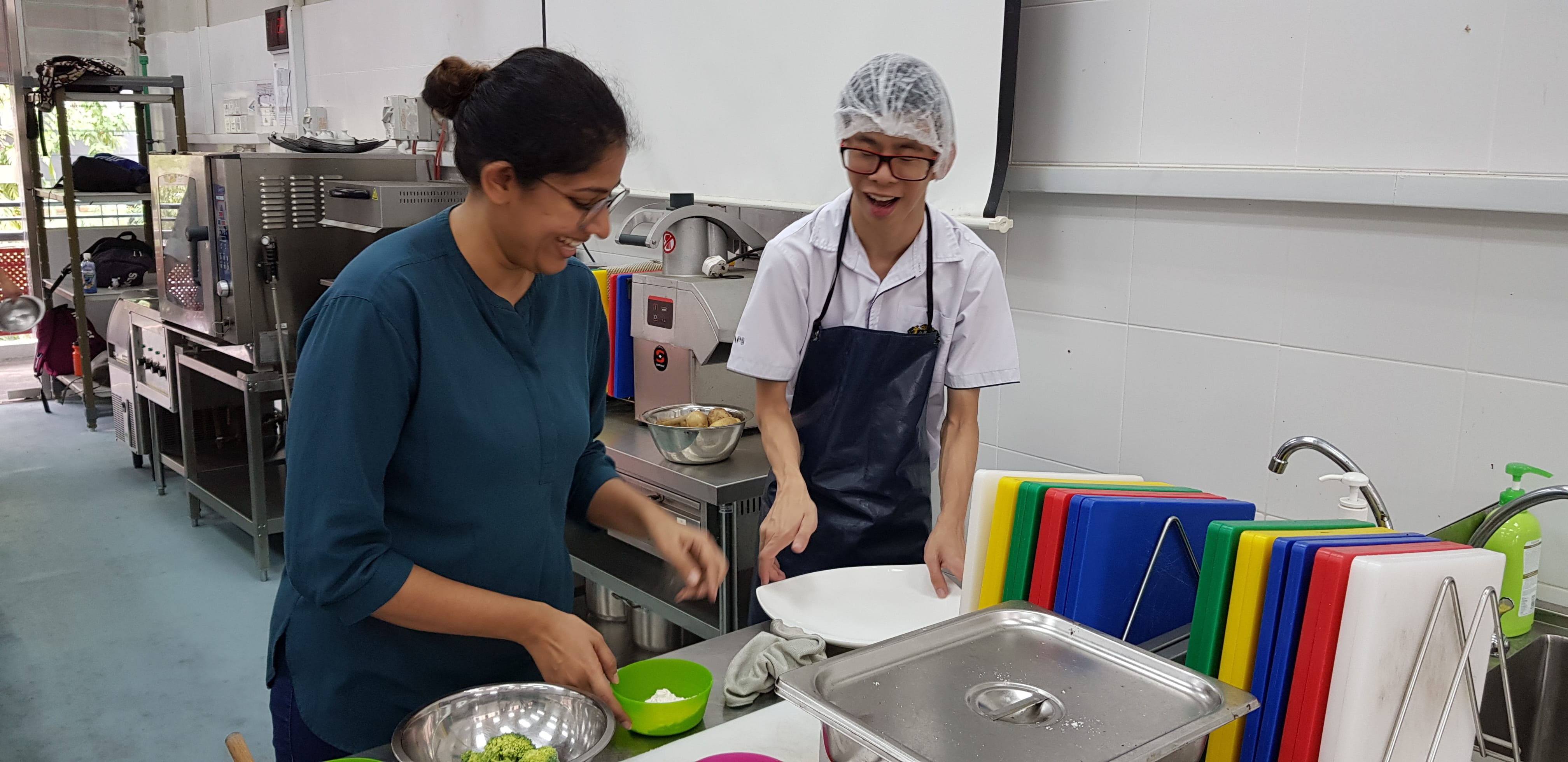Two people smiling while working together in a commercial kitchen with cutting boards, bowls, and stainless steel equipment.
