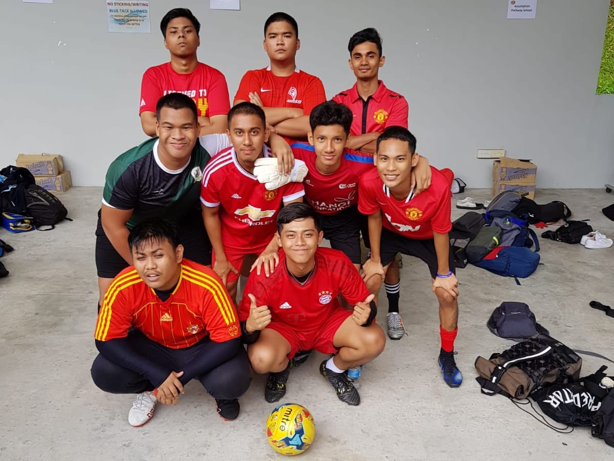 Nine young male football players in red jerseys posing together indoors with a Mitre ball on the floor.