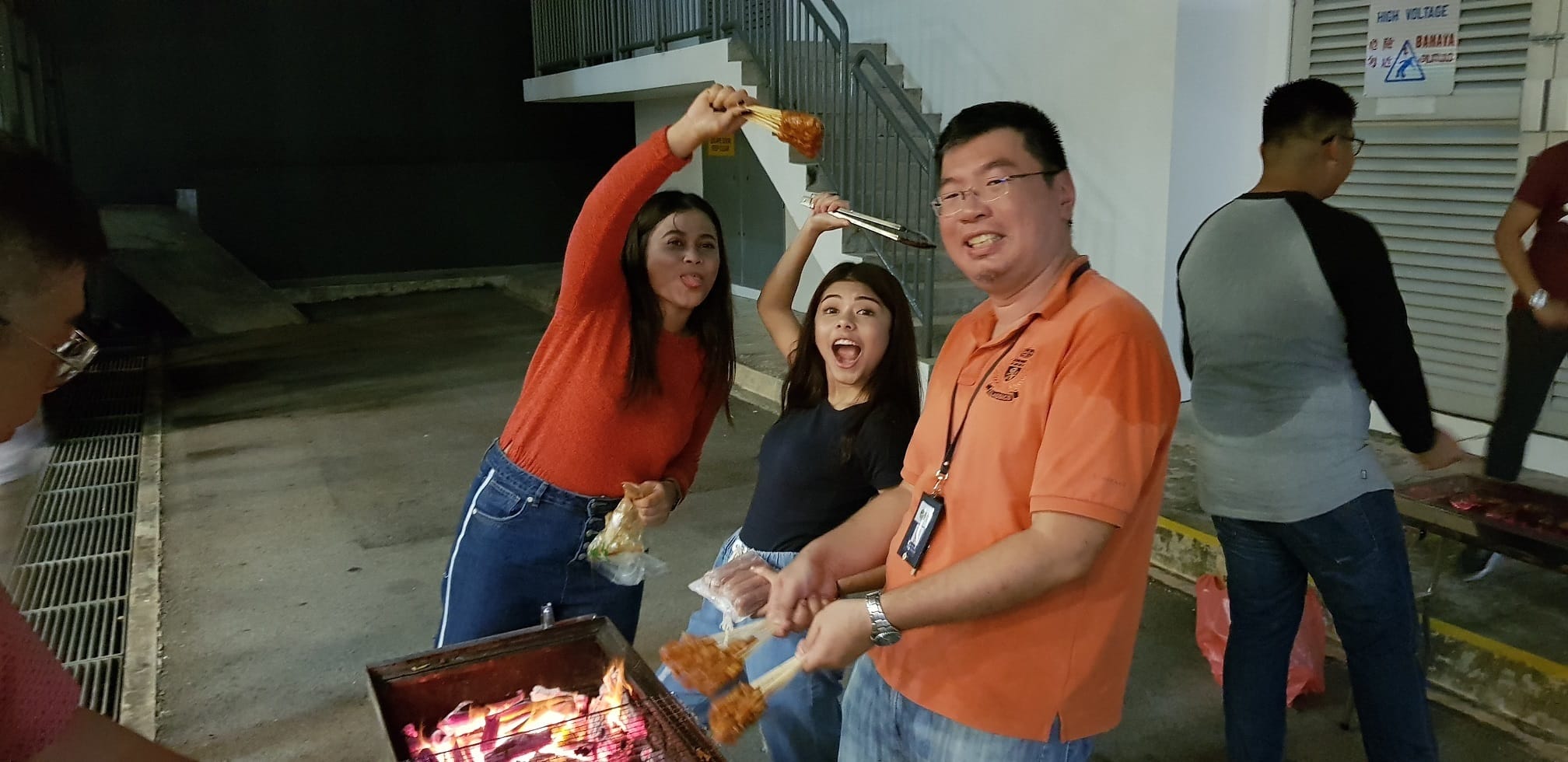 A man and two women posing playfully at a barbecue pit at night, one woman holding up a skewer of grilled food, with other people in the background