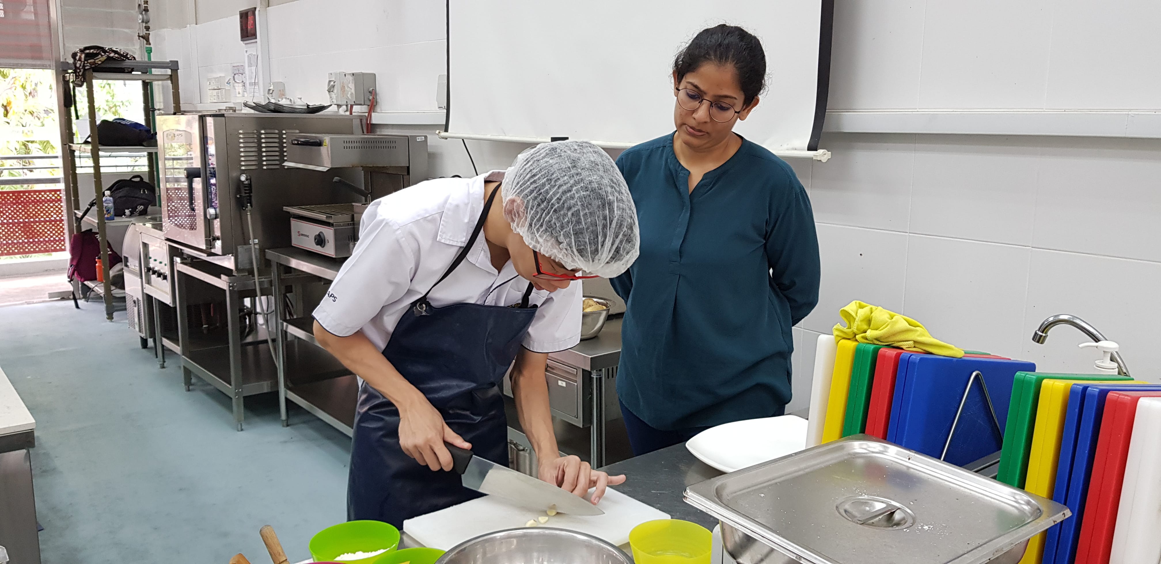 A person in a hairnet and apron chops food on a cutting board while another person watches in a commercial kitchen.