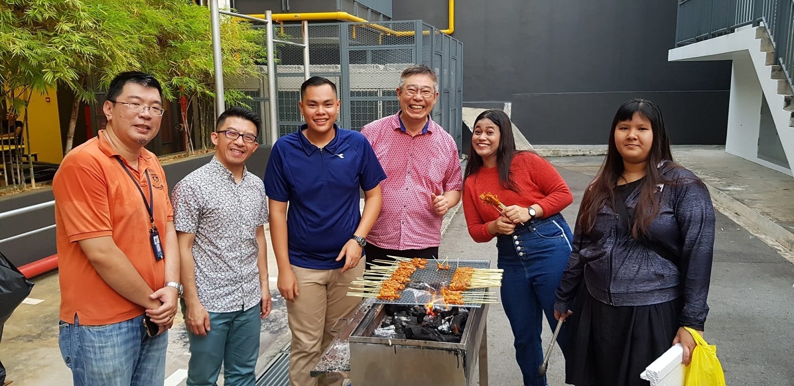 A group of six people, four men and two women, posing together outdoors near a barbecue grill with satay skewers cooking.
