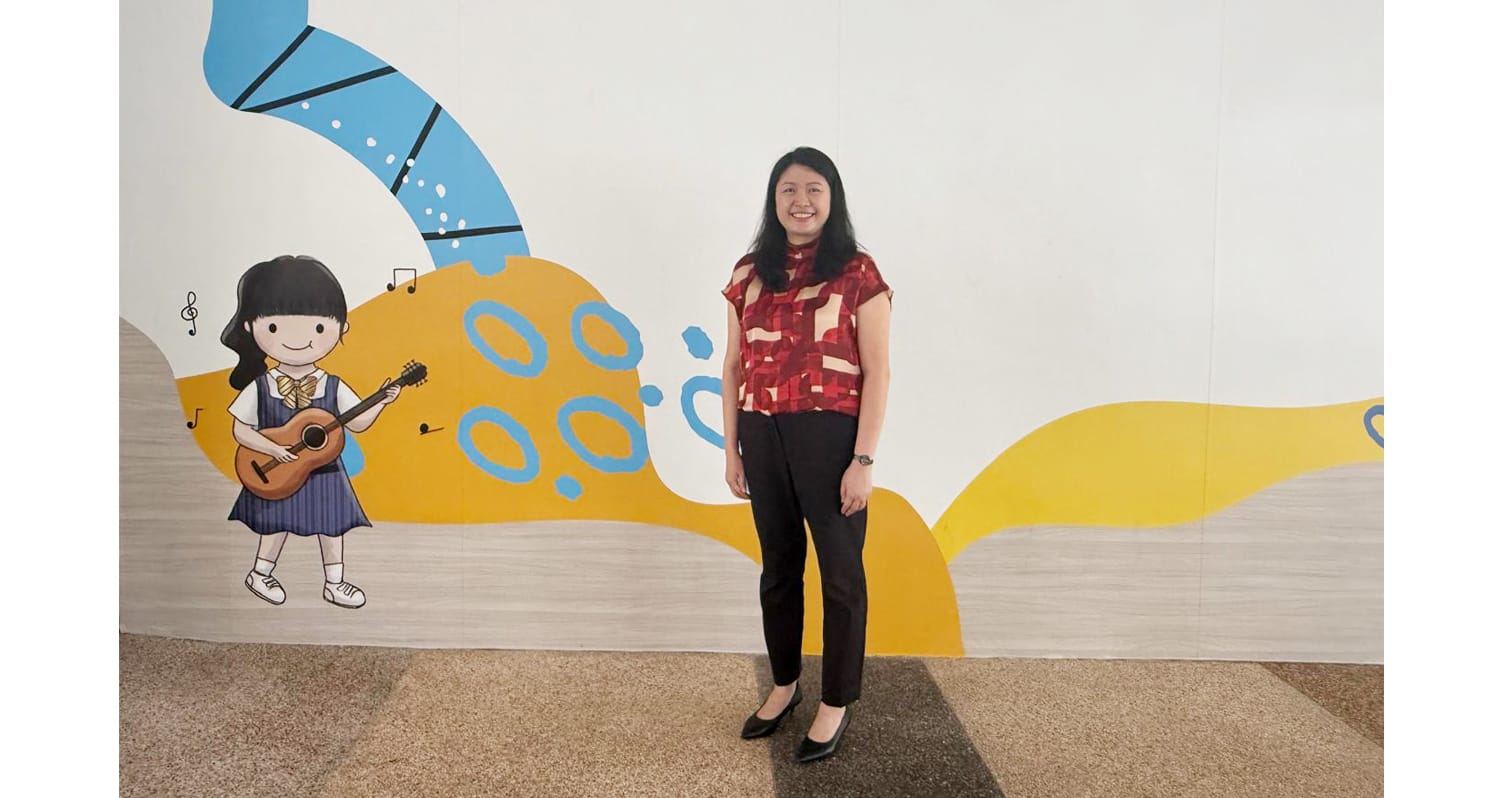 A smiling woman in a red top and black pants stands before a colorful mural showing a cartoon girl playing guitar.