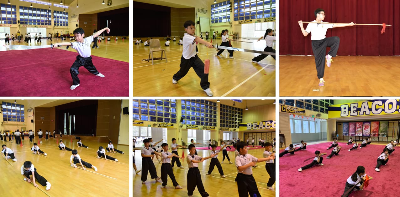 Students in black and white uniforms practicing wushu martial arts with staffs and swords in a school gymnasium.