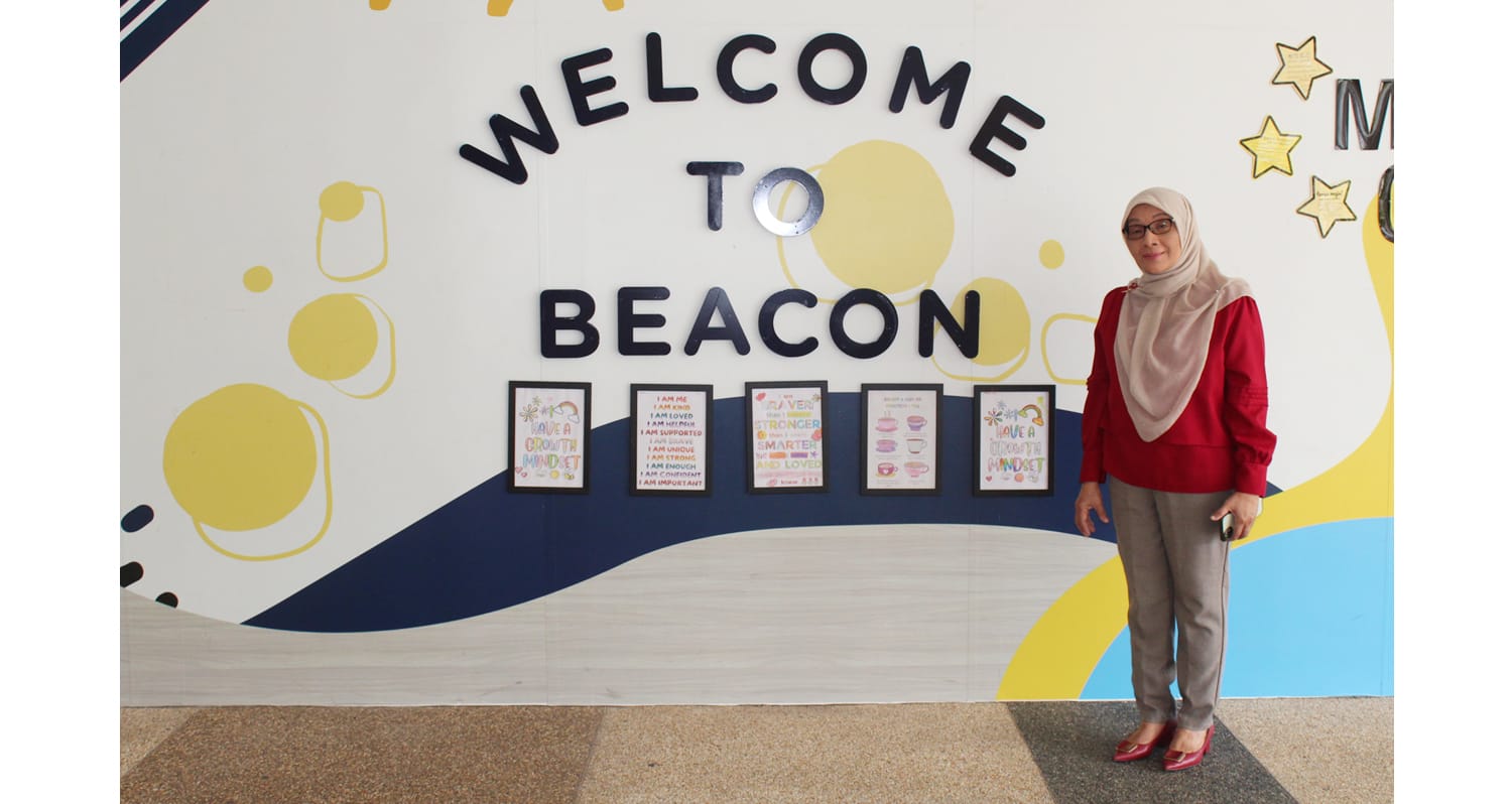 Woman in red top and hijab standing in front of a colorful "Welcome to Beacon" wall with framed posters.