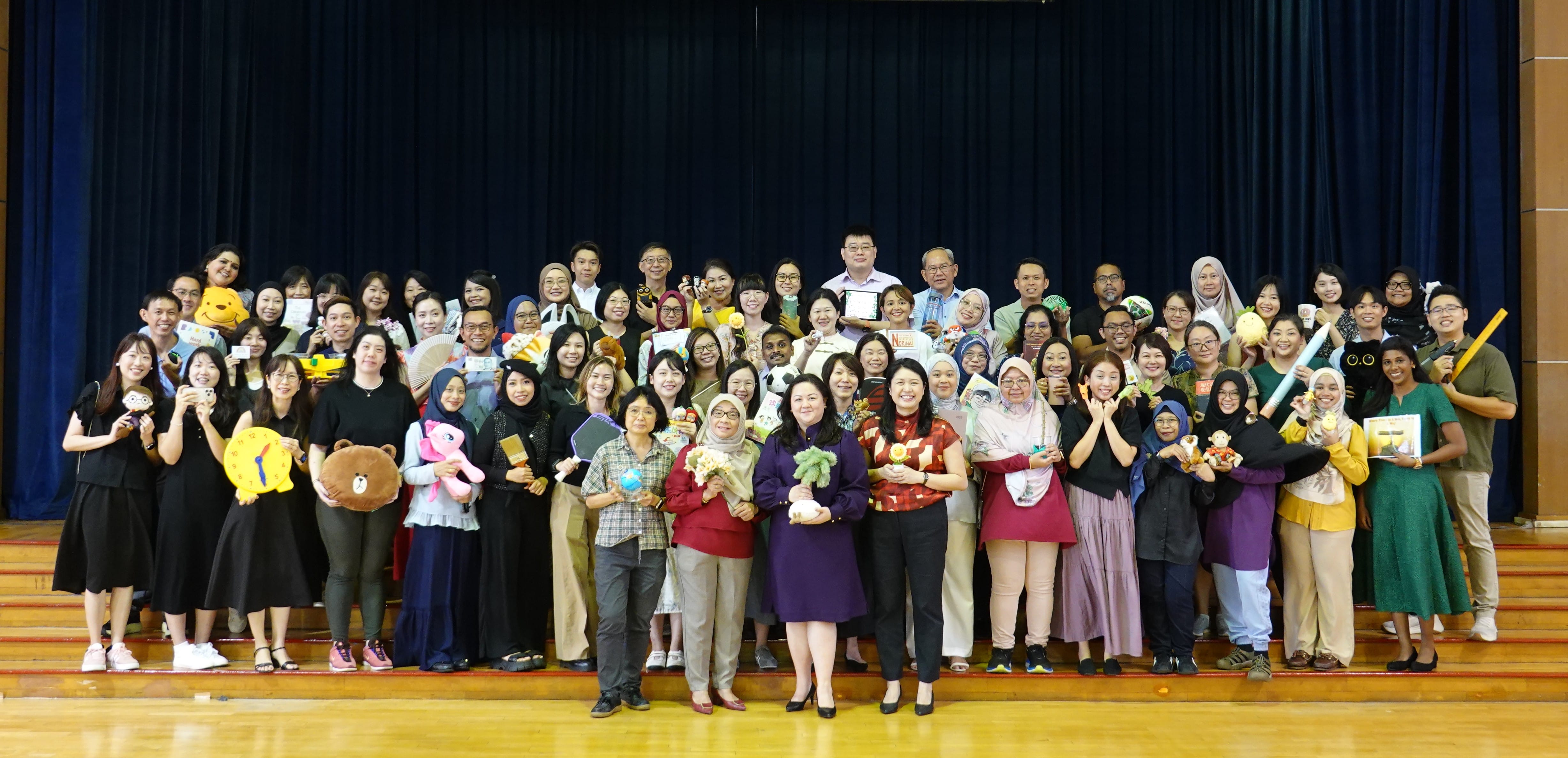 Large group of adults posing on a stage, holding stuffed toys, books, and colorful items, smiling at the camera.
