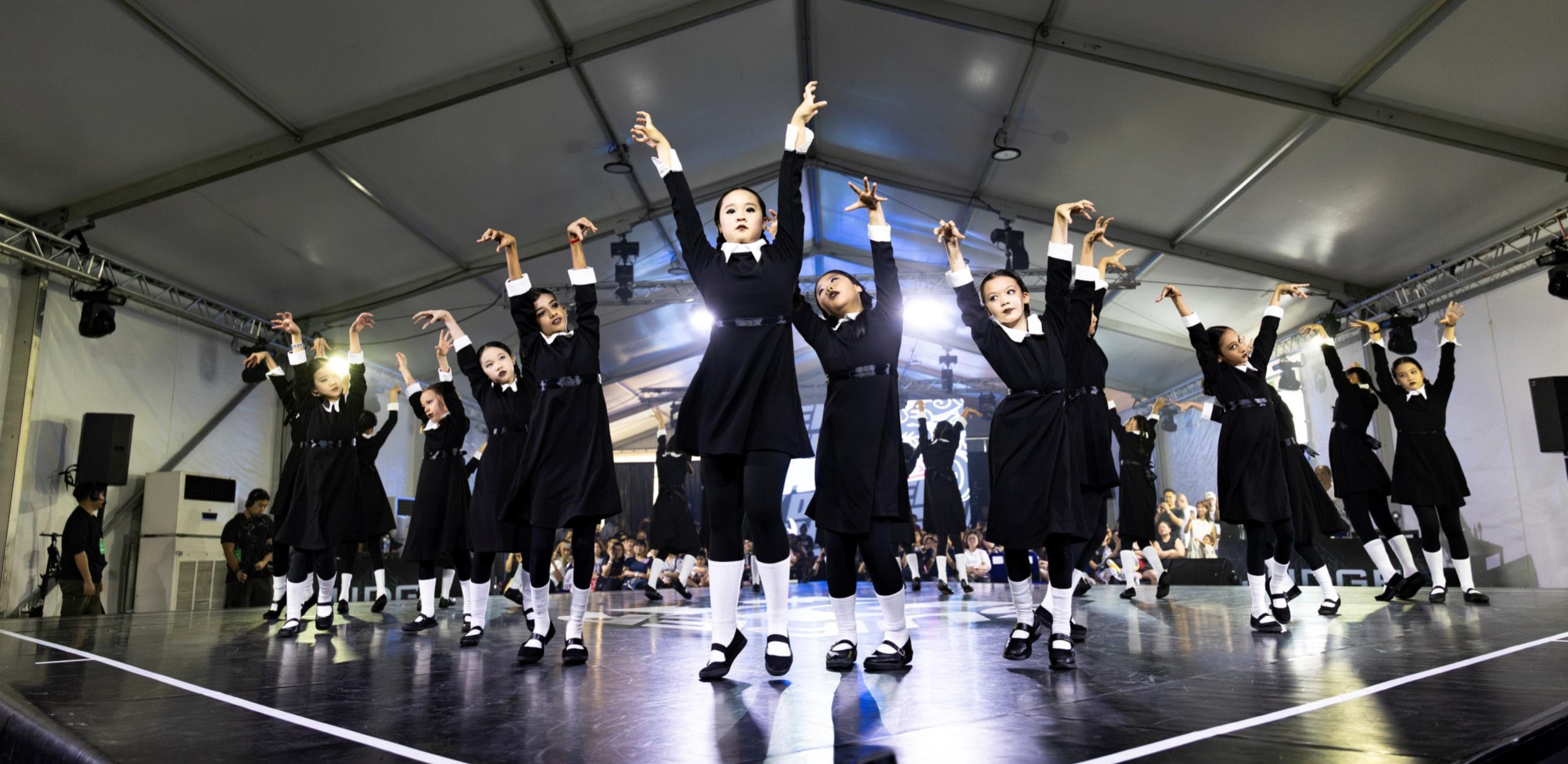 Students in black costumes perform a choreographed dance routine on stage under bright lights.
