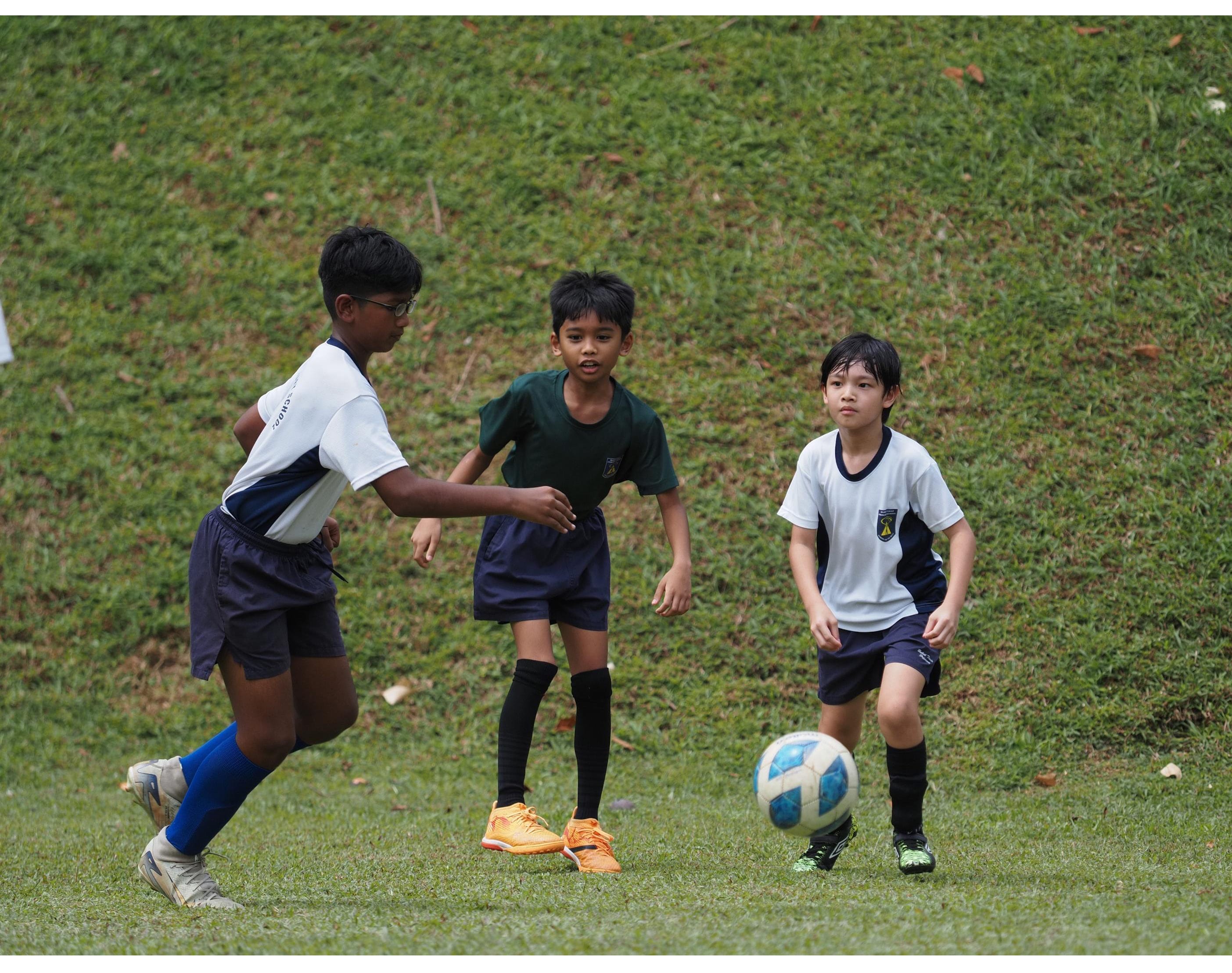 Three boys in sports uniforms play football on a grass field, with one kicking a blue and white ball.