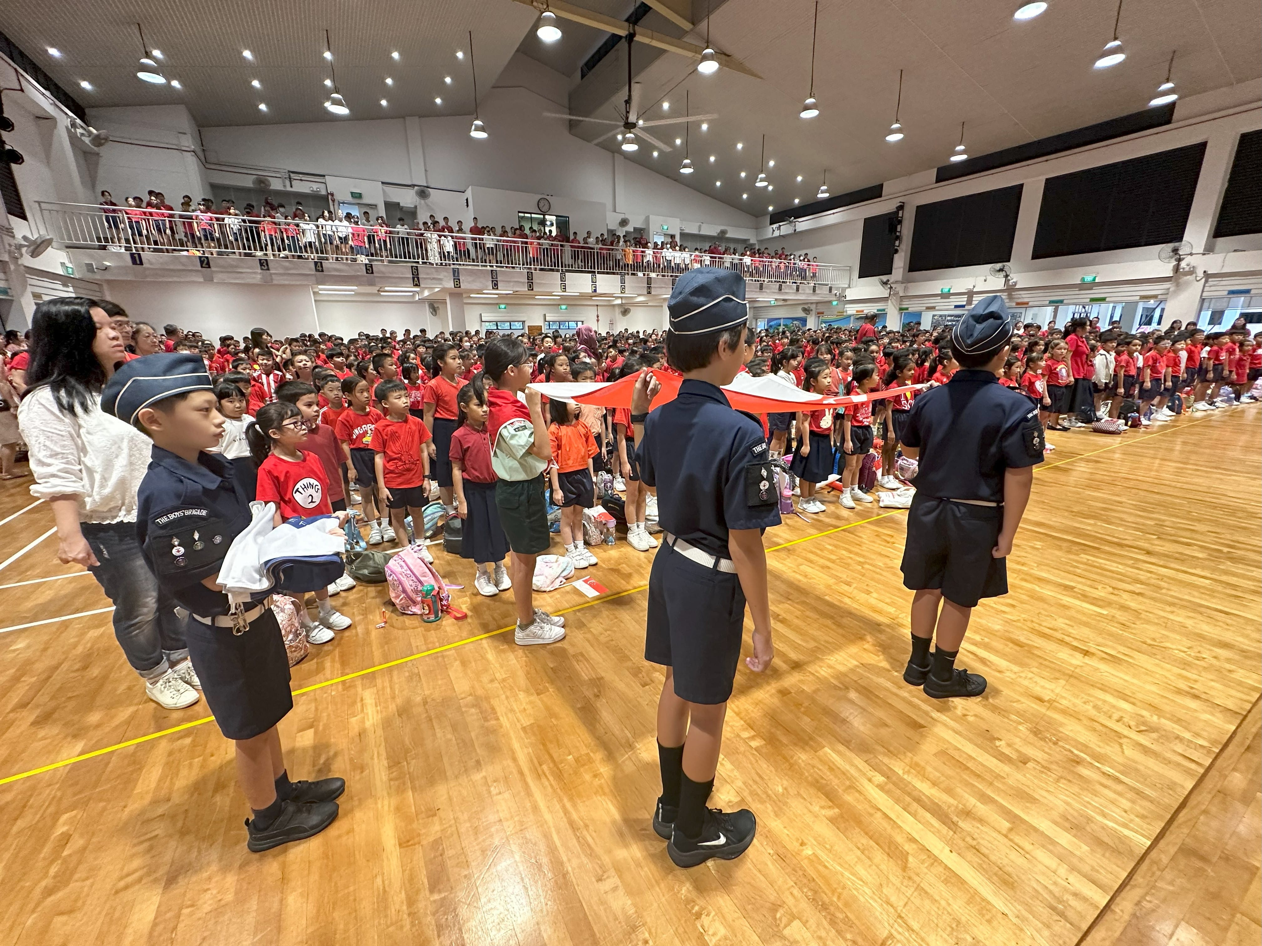 Uniformed student leaders carry flags during a ceremony in a packed school hall, with students on the floor and upper gallery.