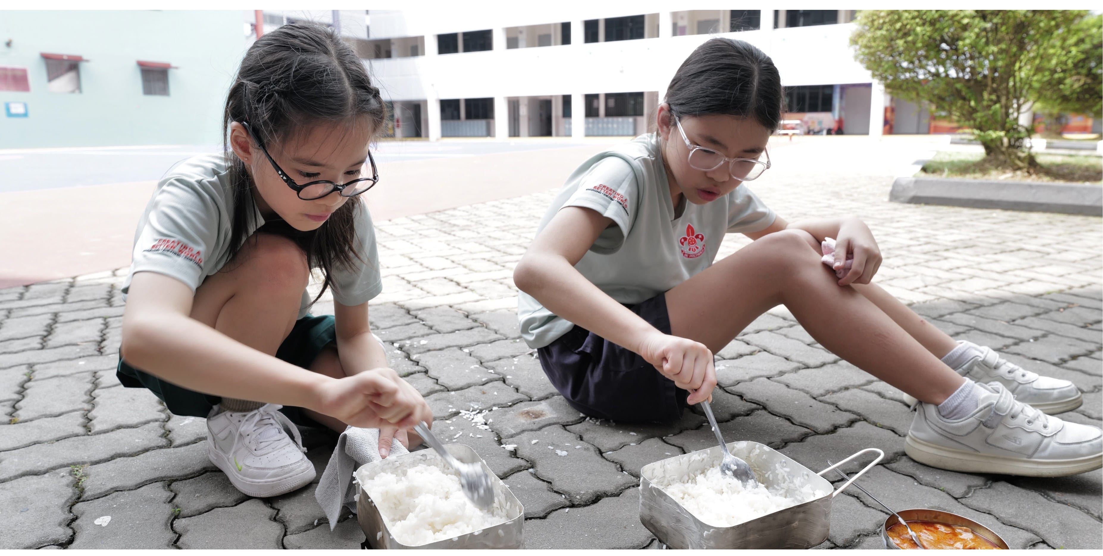 Two students mix food ingredients in metal containers during an outdoor activity in the school courtyard.