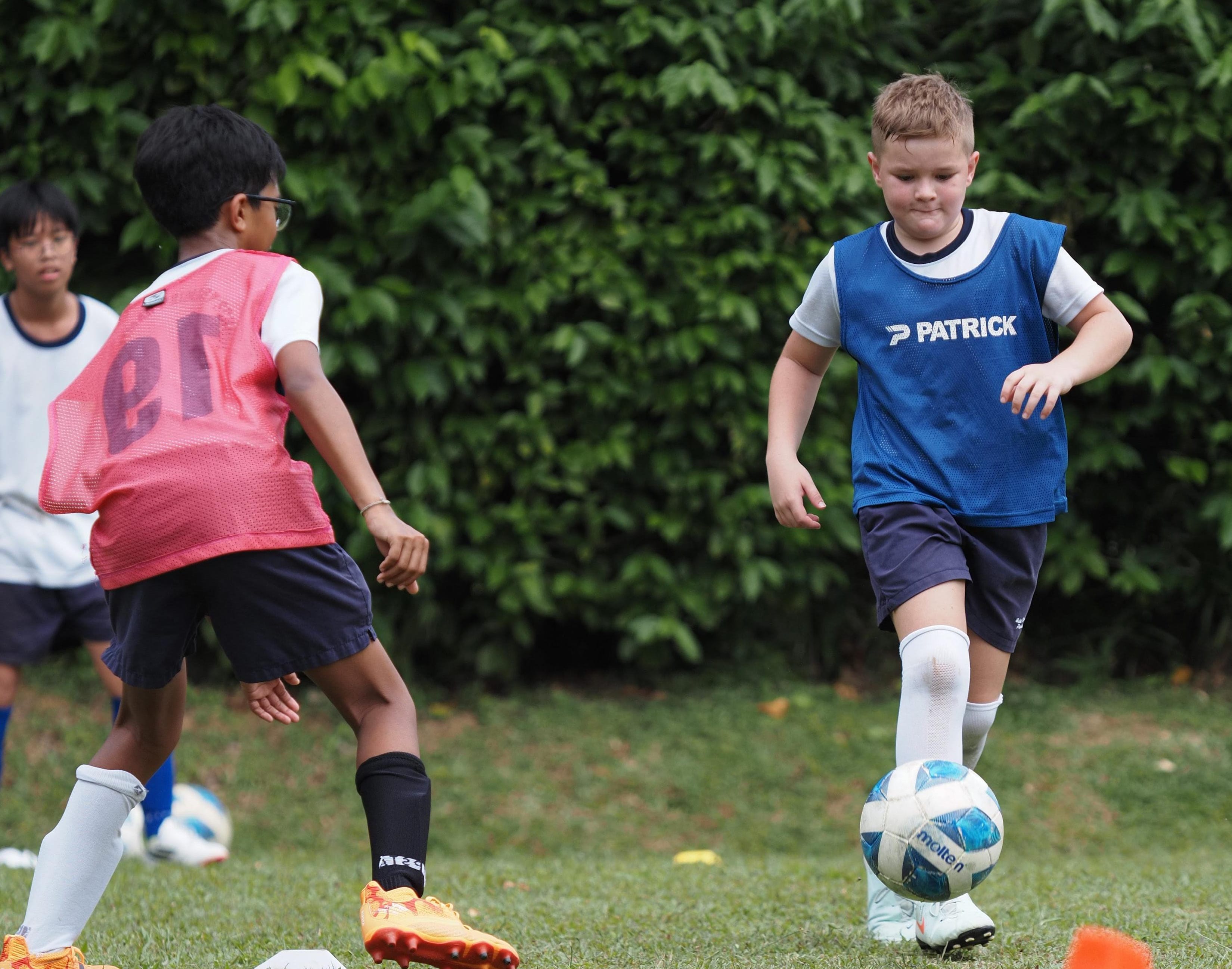 Two boys in coloured vests play football on grass, one dribbling towards the other.