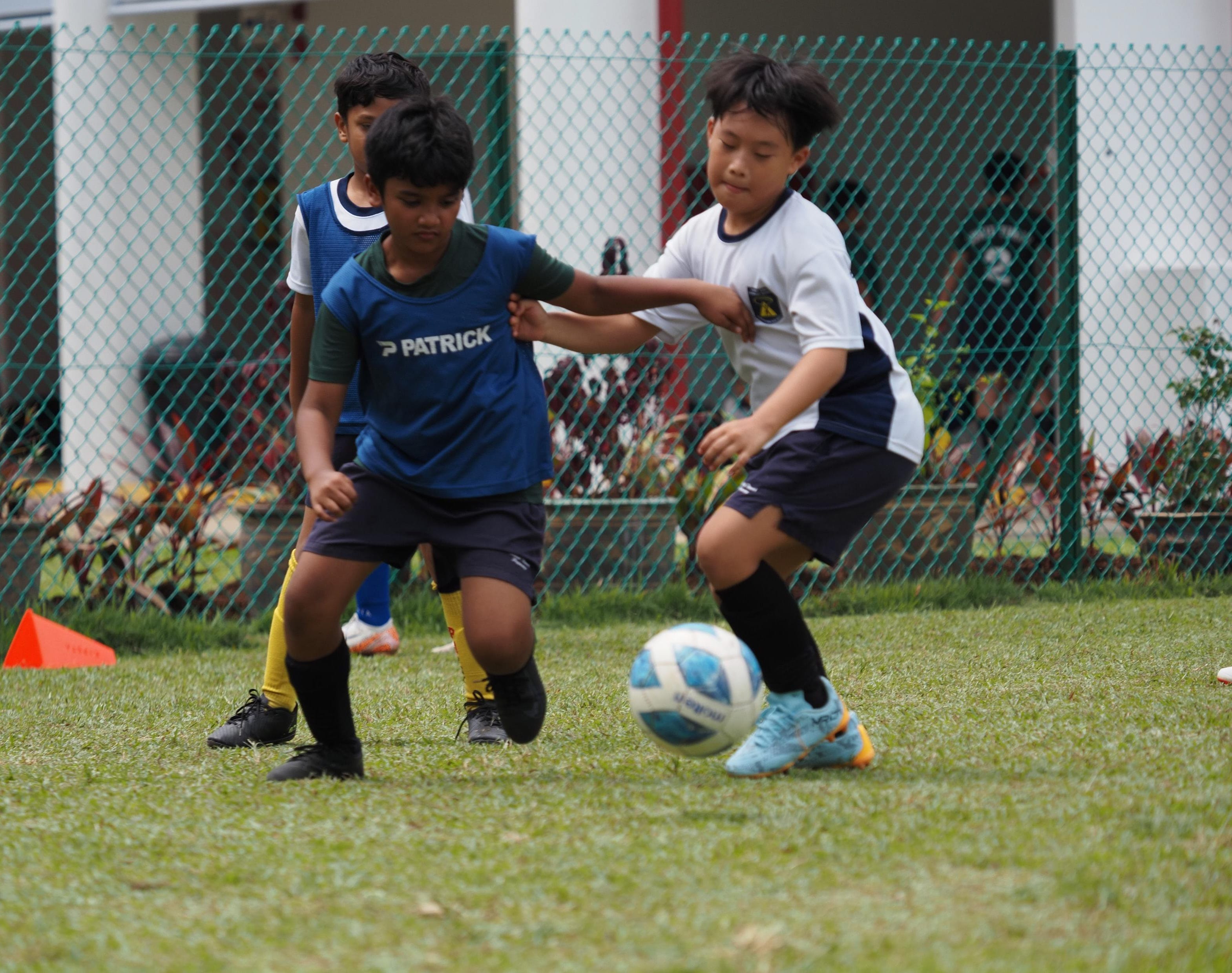 Two young boys compete for a football on a grass field, with a third boy and a chain-link fence behind them.