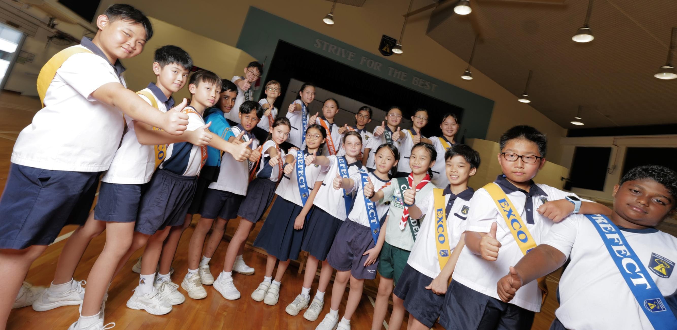 Student leaders in "EXCO" and "PREFECT" sashes give thumbs-up in the school hall.