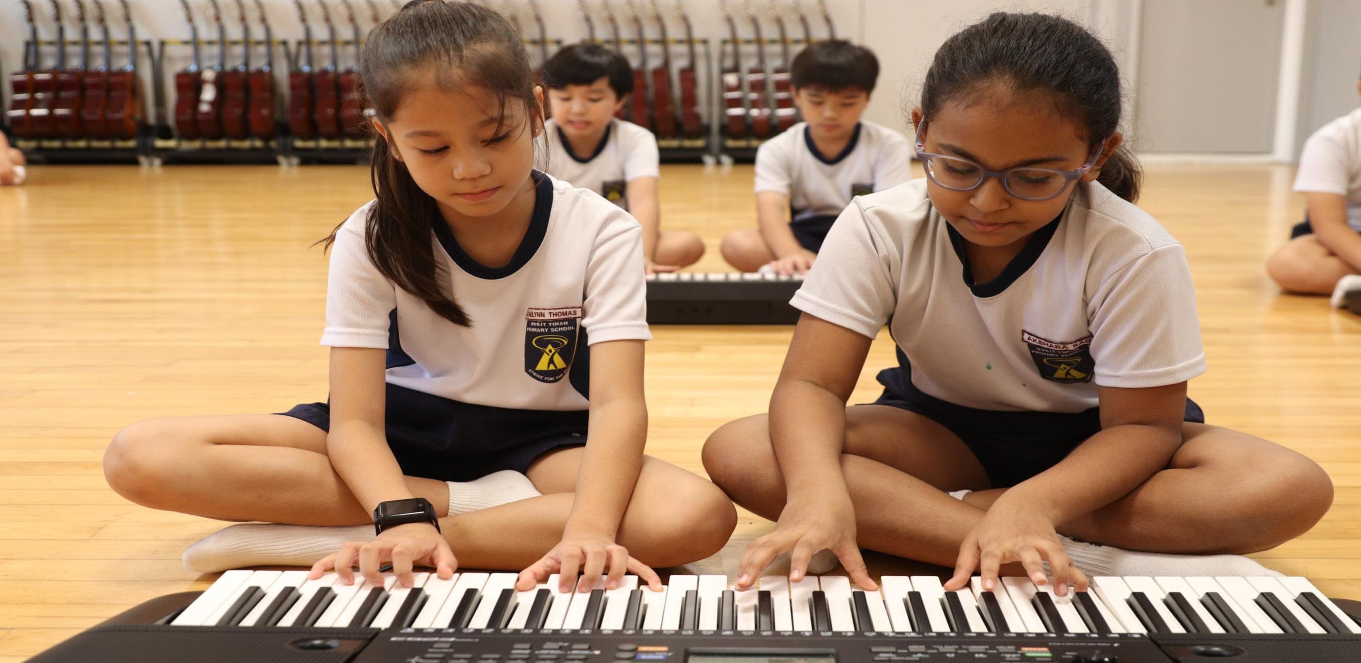Two students collaborate on a keyboard during a music lesson, with classmates practising in the background.