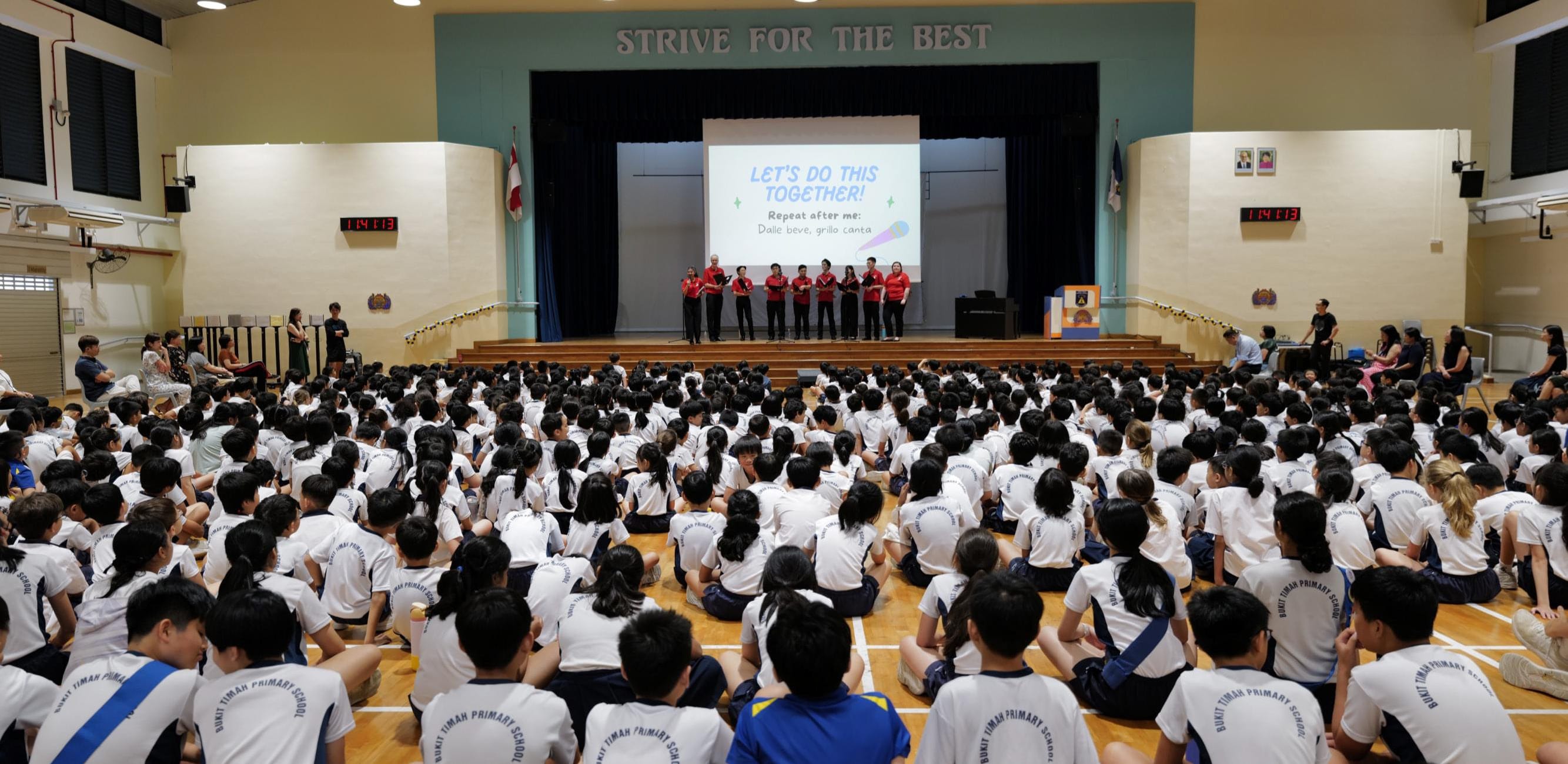 Staff lead an assembly presentation on stage, with students facing a screen displaying "Let's Do This Together!"