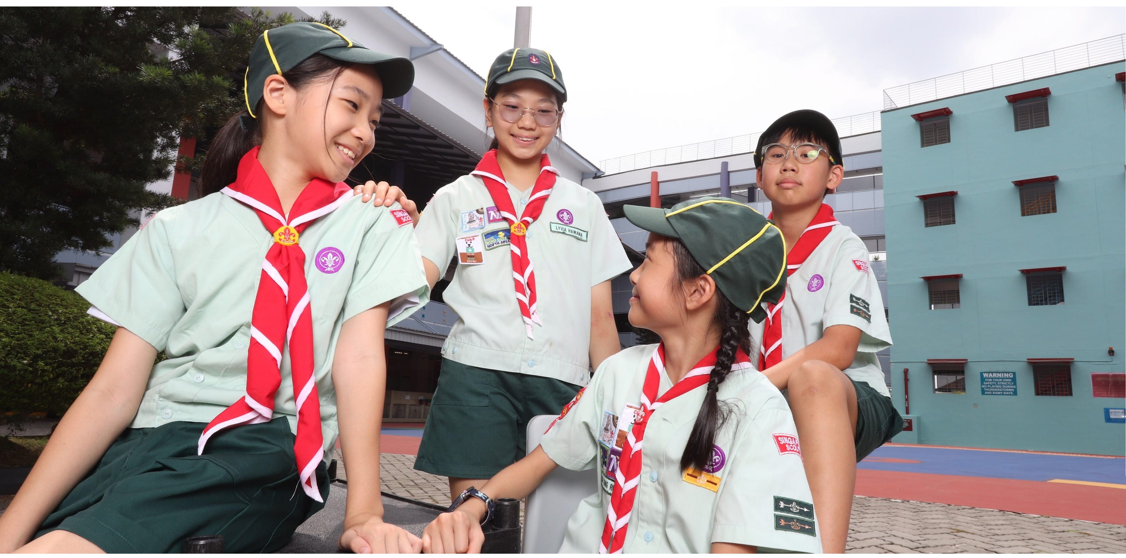 Four students in scout uniforms gather in a school courtyard.