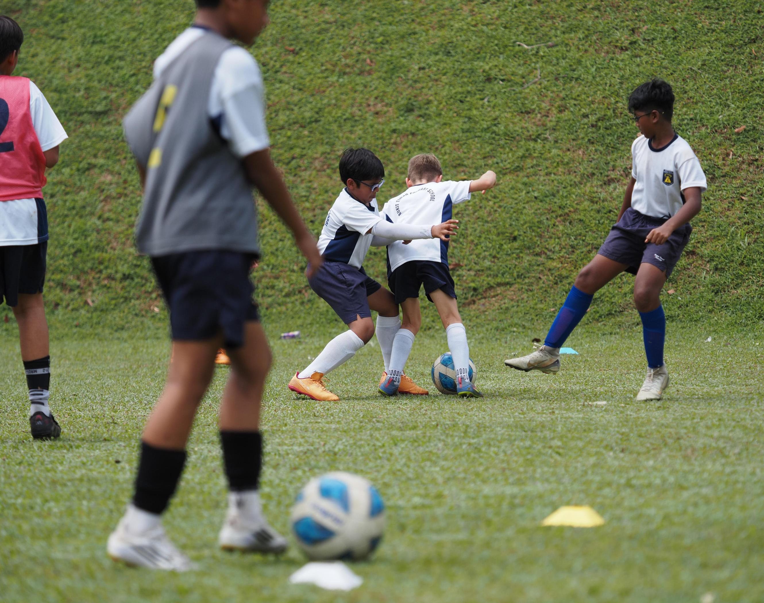 Several young boys in white and dark shorts compete for a football on a green grass field.