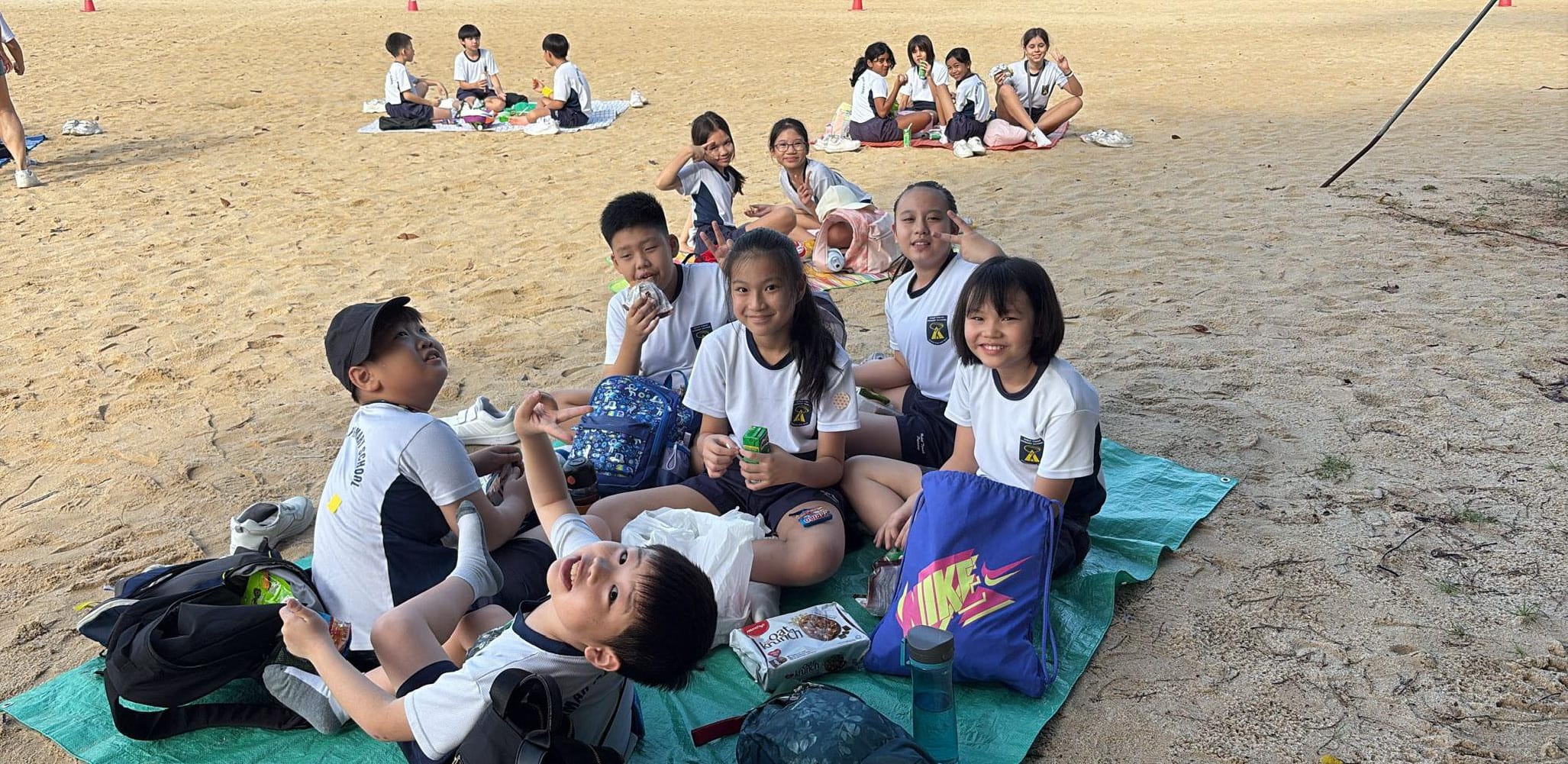 Students relax on picnic mats on a sandy field, enjoying snacks and socialising during a break.
