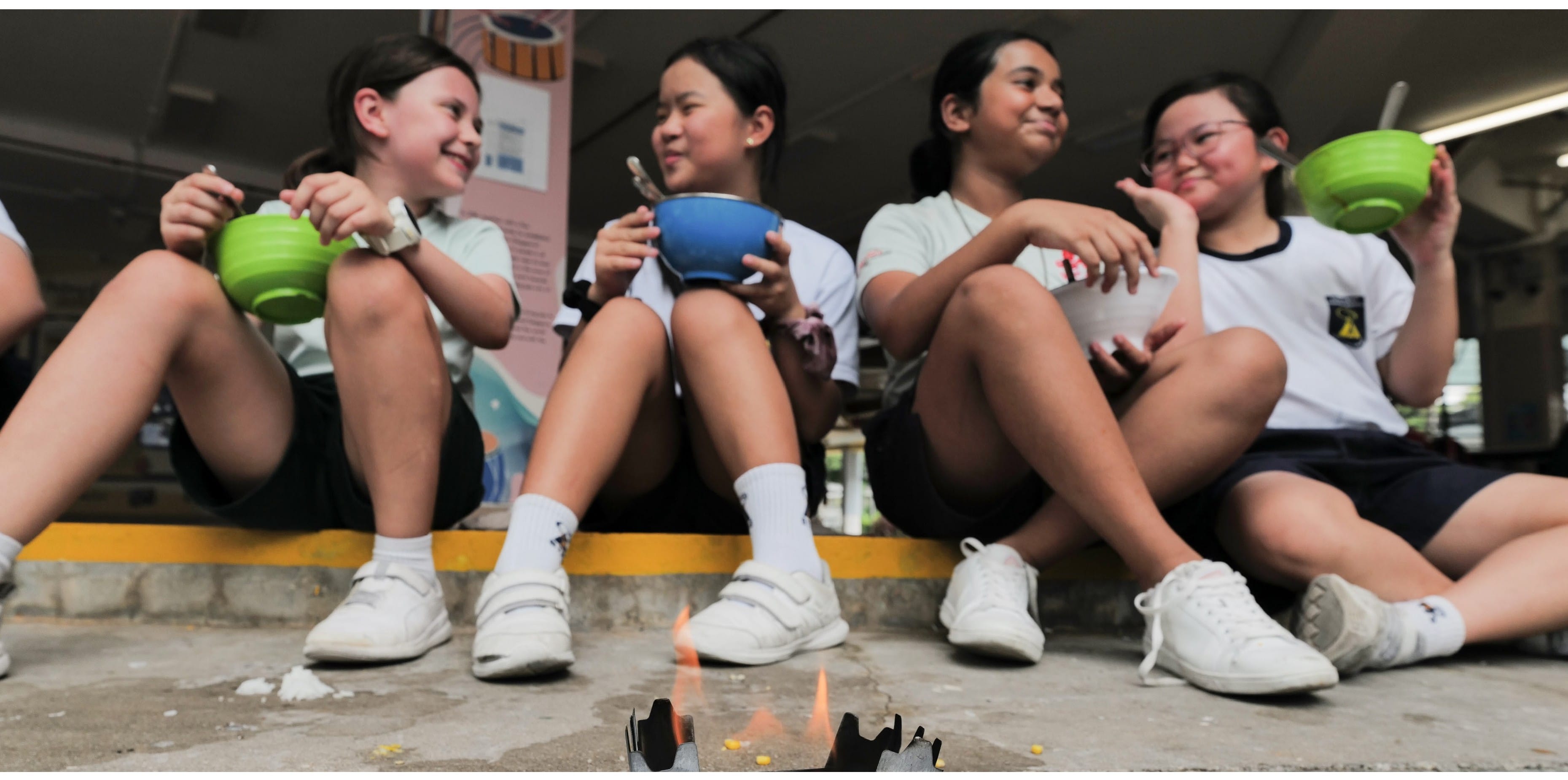 Four students sit on a school step with bowls and spoons gathered around a small campfire.