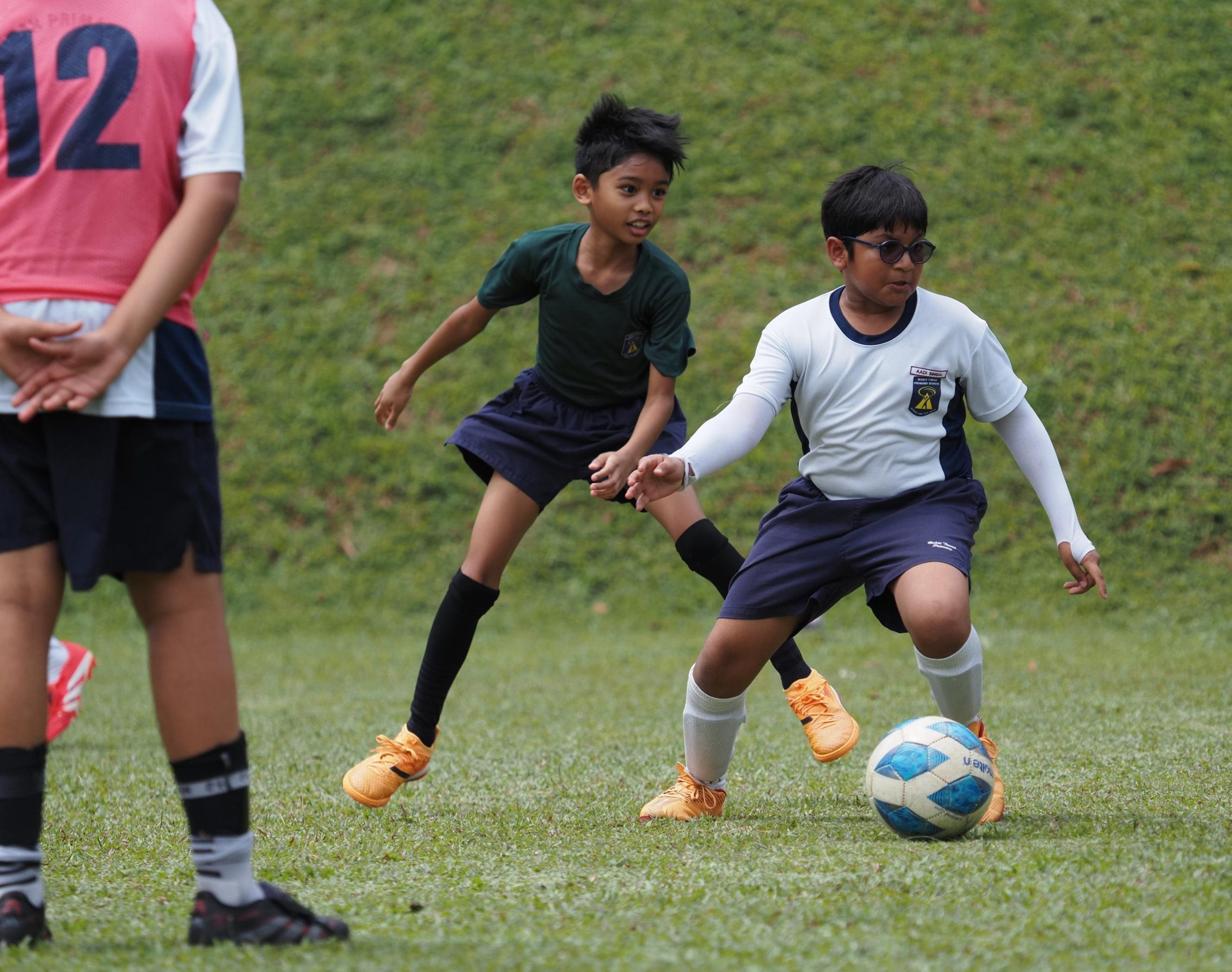 Three young boys playing football on a grass field, one dribbling a blue and white ball while another defends.