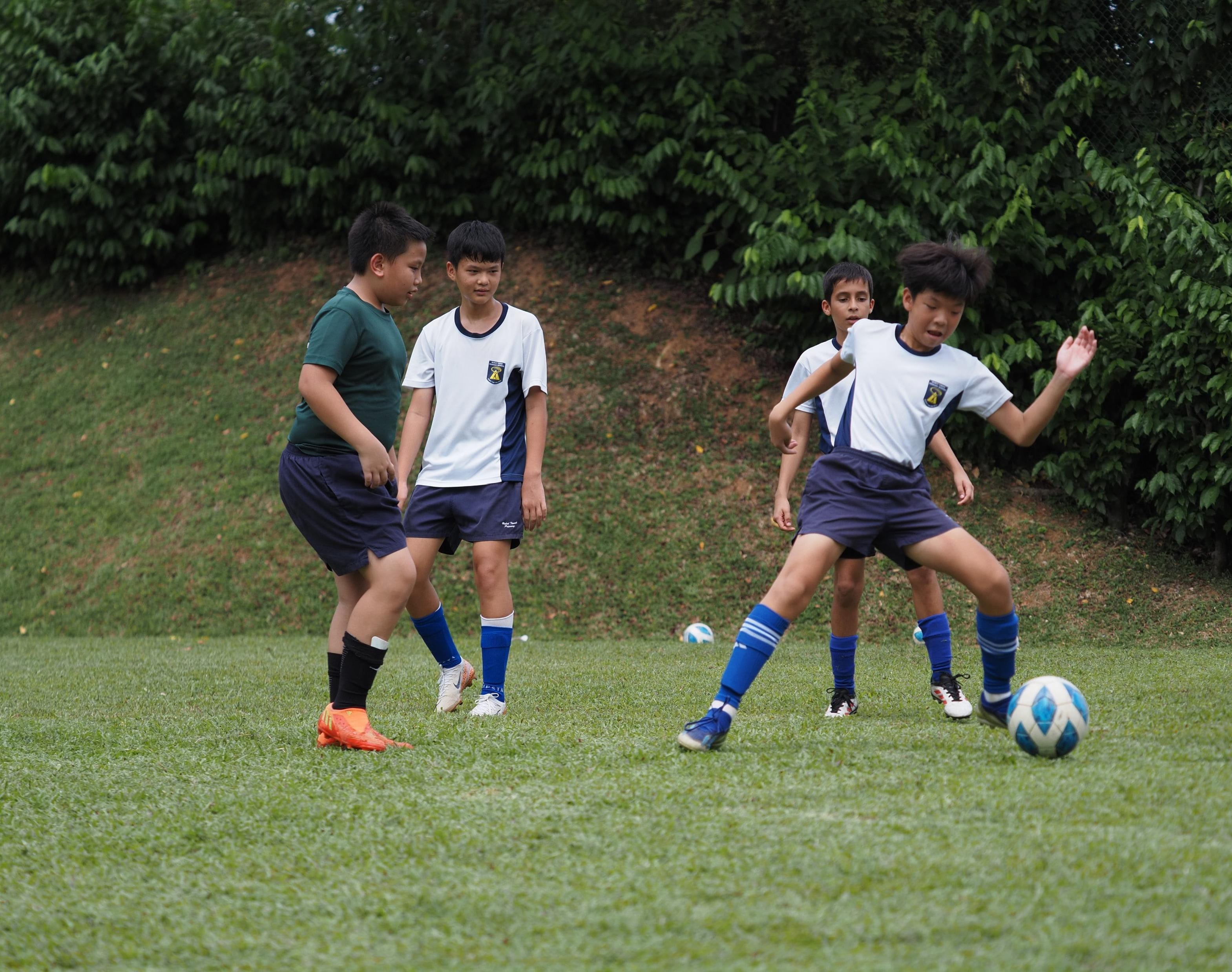 Four boys playing football on a grass field, one about to kick a blue and white ball.