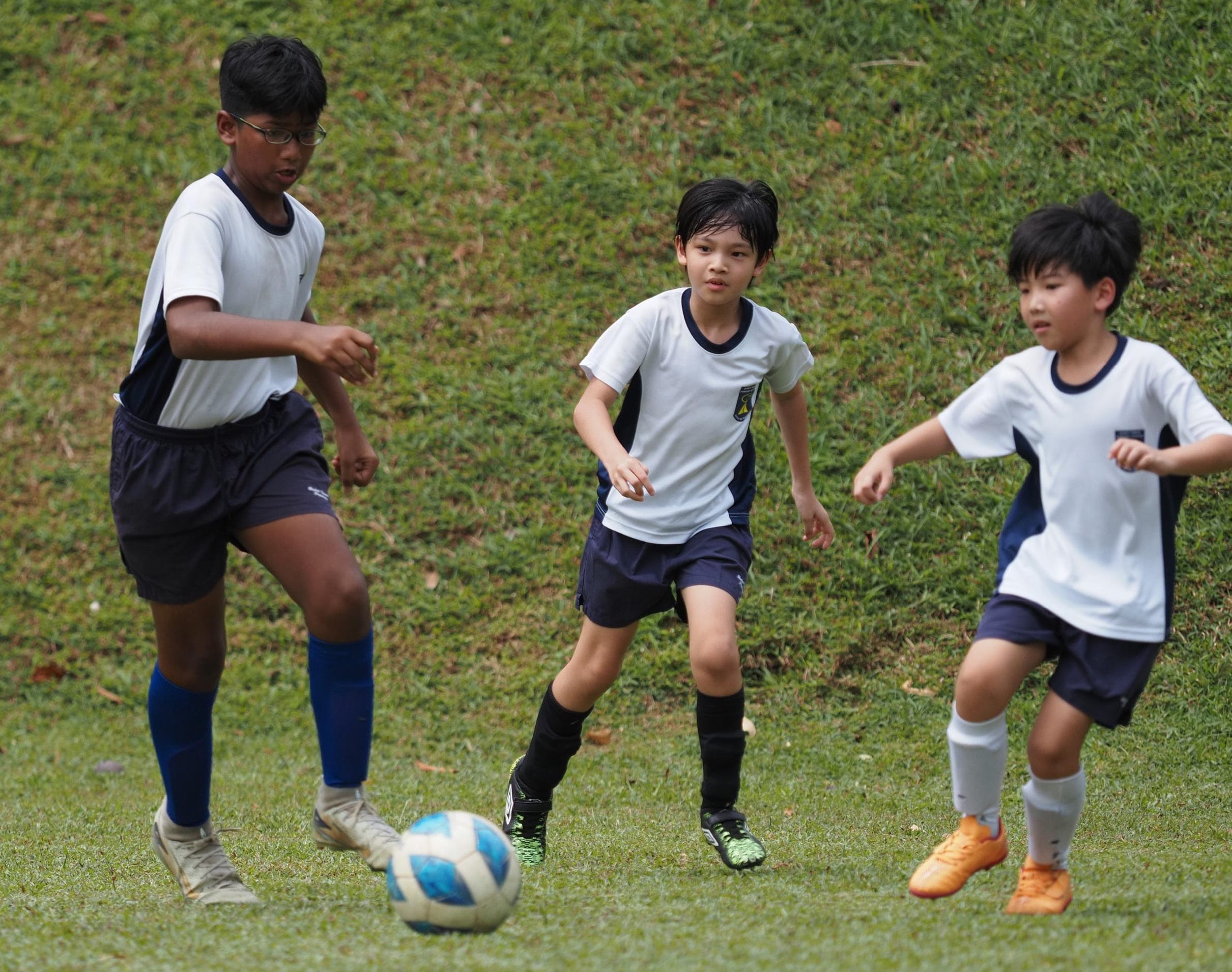 Three boys in white and navy uniforms play football on a grass field, one about to kick the ball.