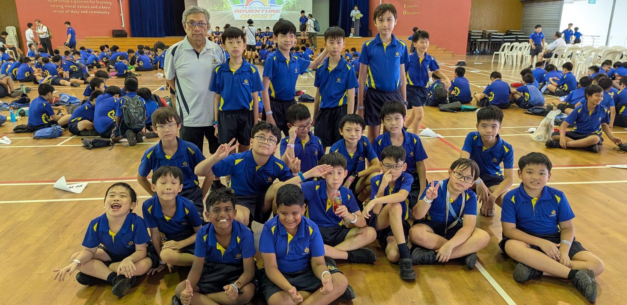 A teacher and students pose for a photo in the school hall, with more students active in the background.