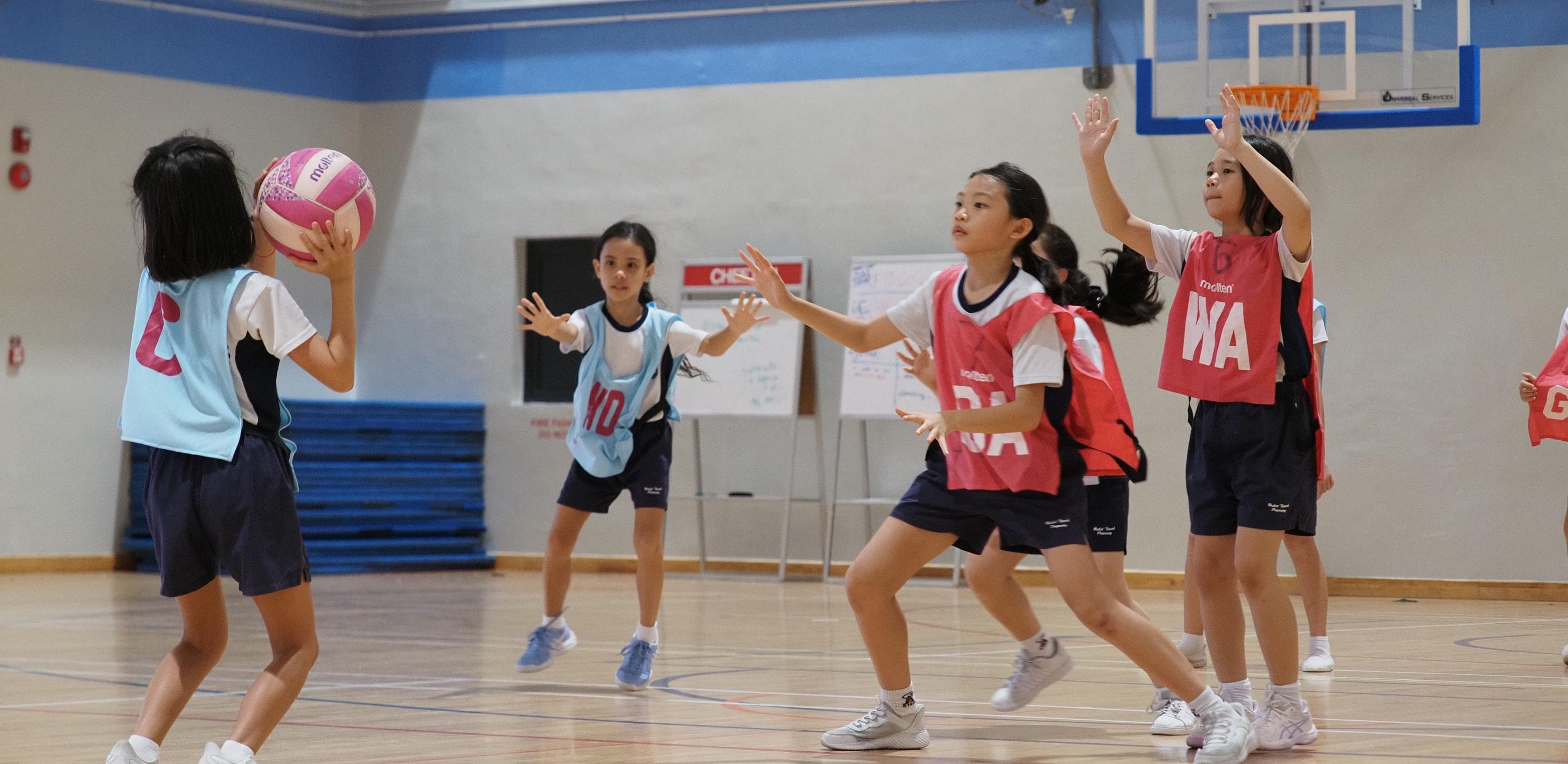 Girls in red and blue bibs play netball indoors, one holding a pink ball near a hoop.