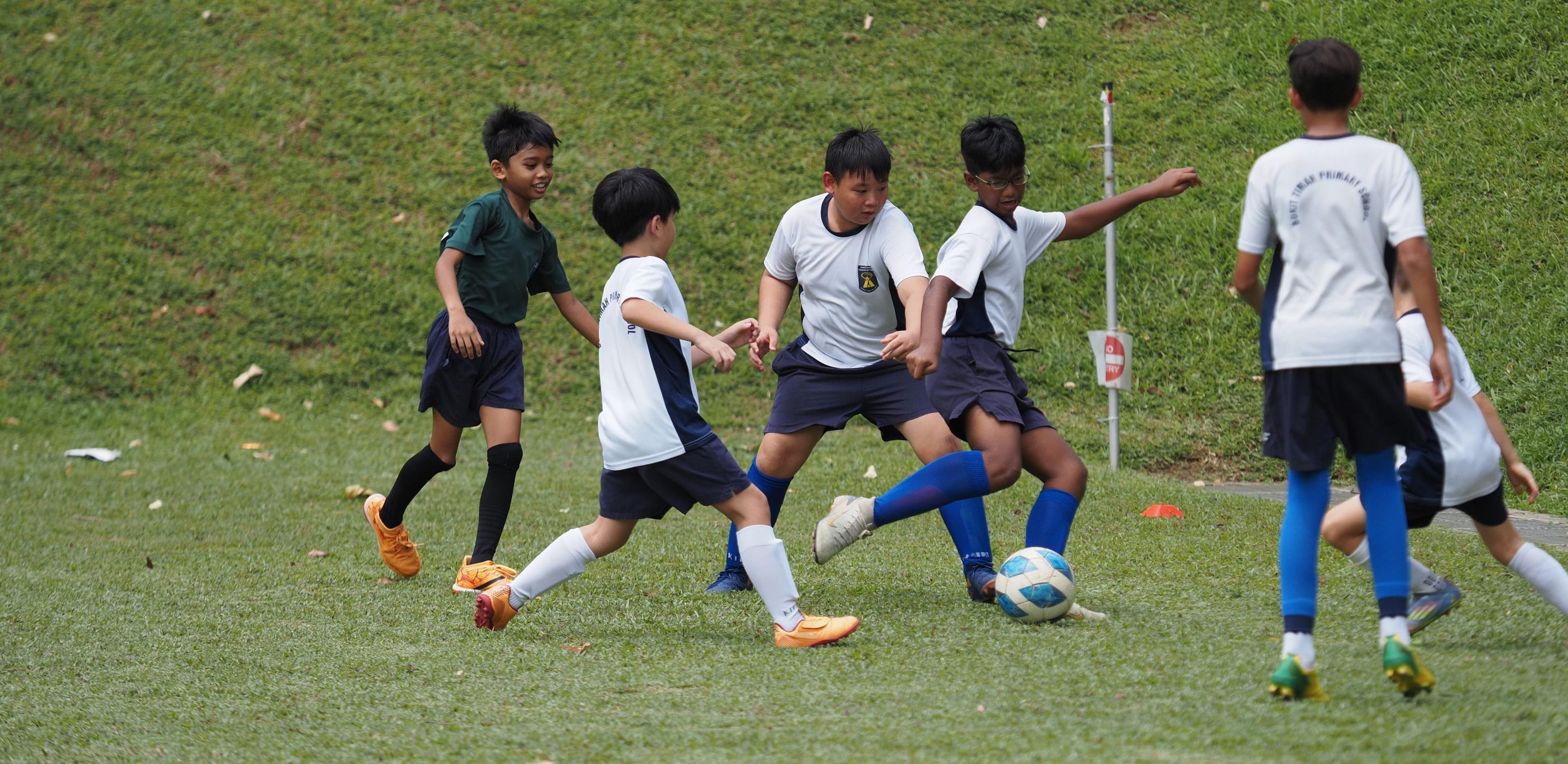 Five boys in white and dark uniforms play football on a grass field, competing for the ball.