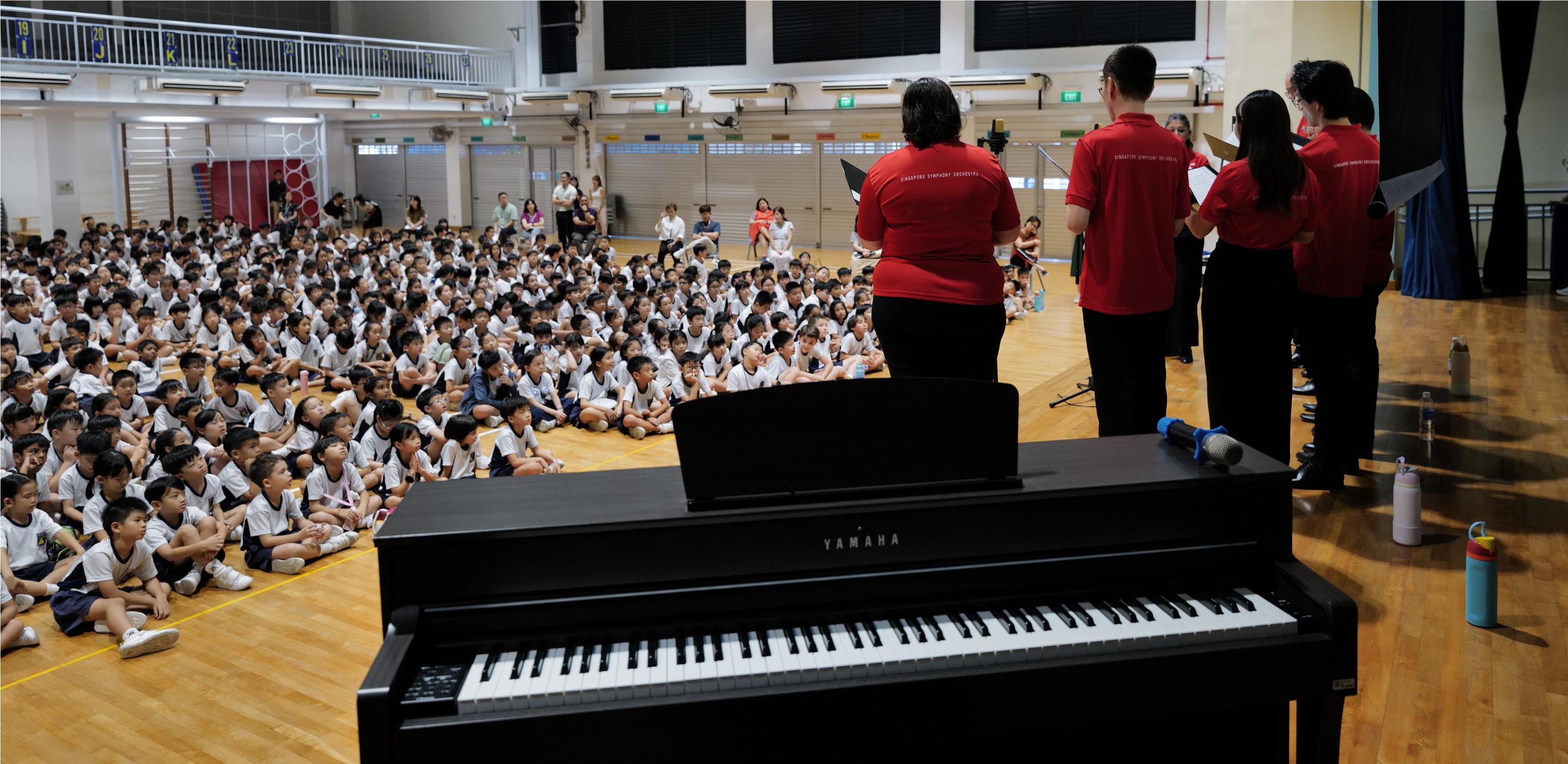  Large audience of students sits attentively on the floor, with a piano visible in the foreground.