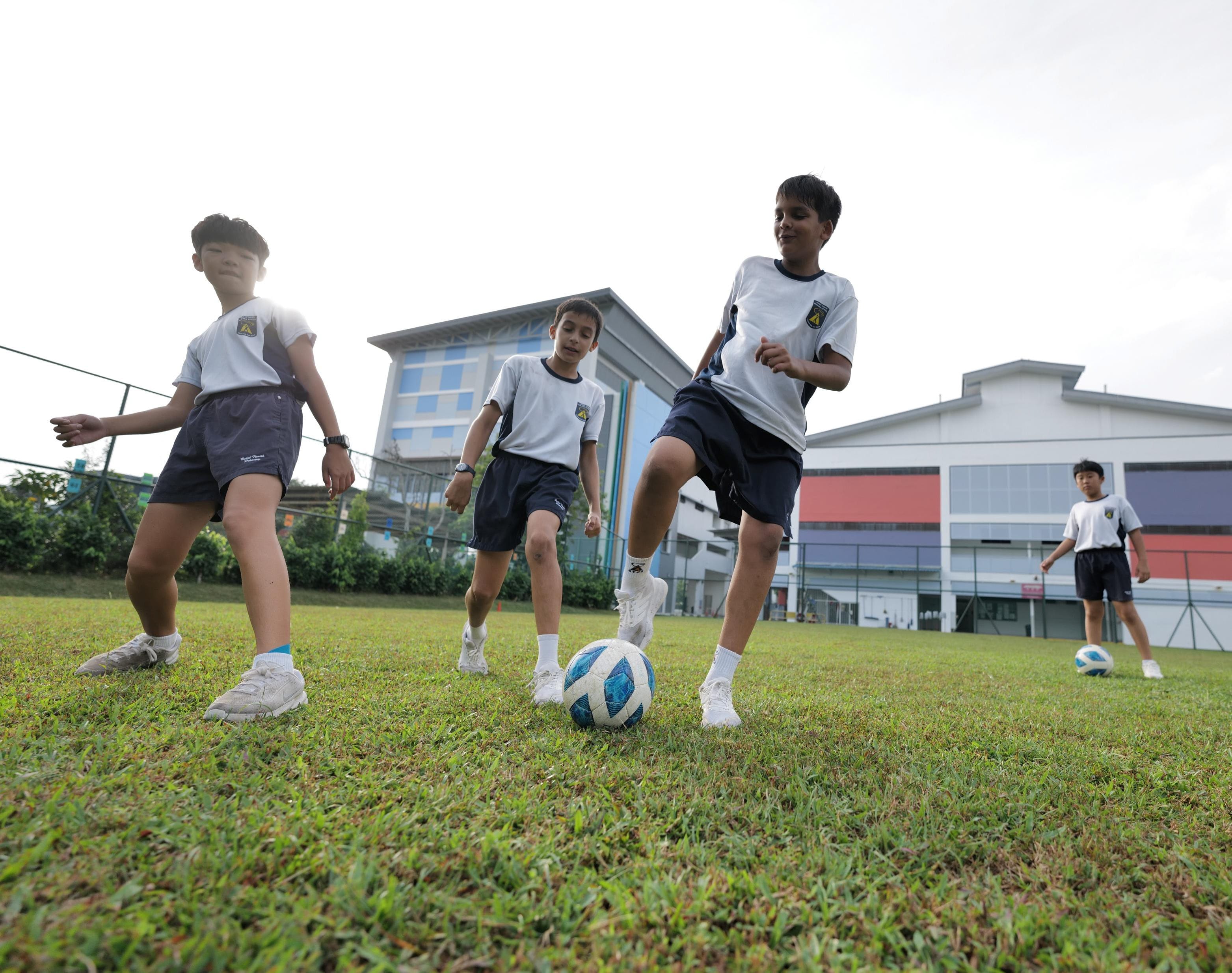 Four children in white and navy school uniforms play football on a grass field.