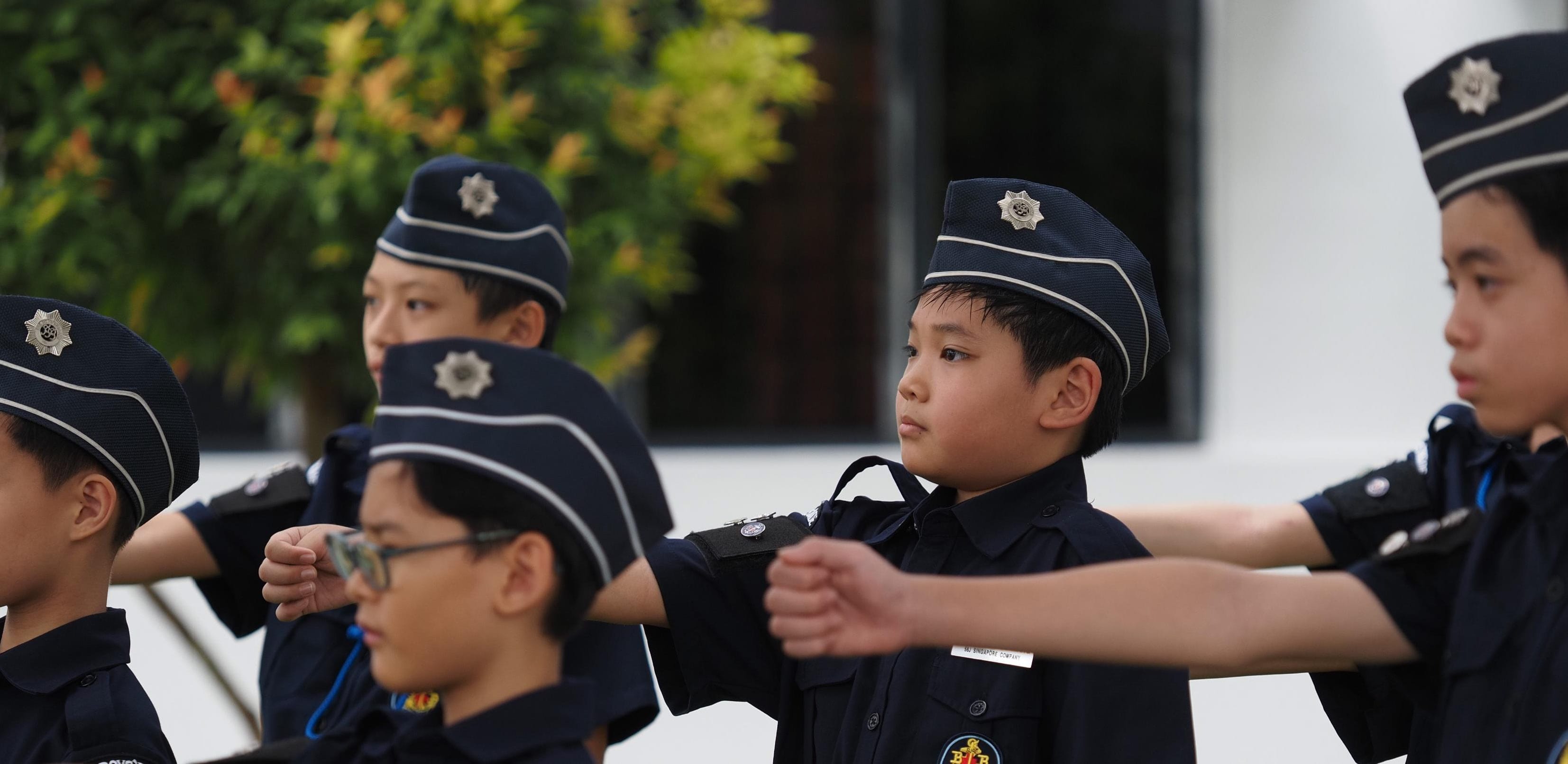 Students in blue uniforms sit together in the school courtyard holding sunflower crafts.