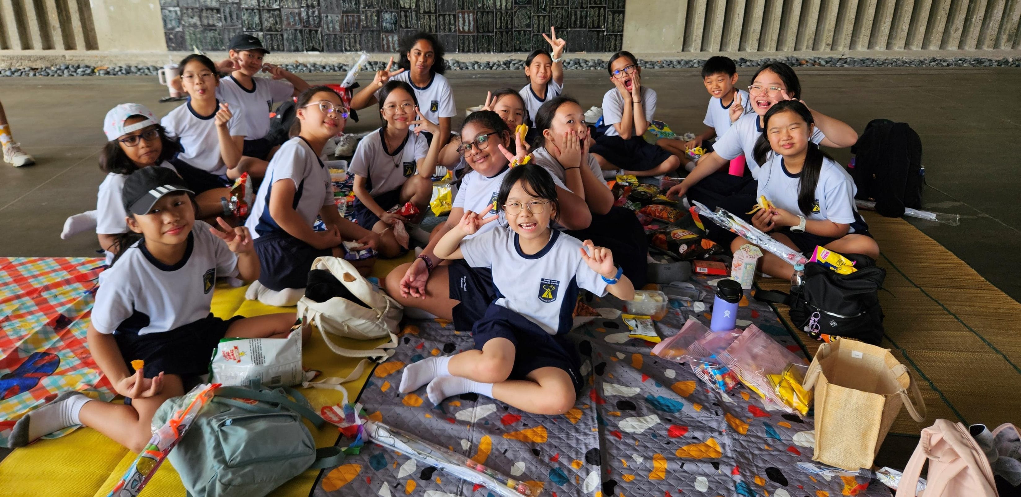 Students relax on picnic mats, sharing snacks during a group break at a sheltered outdoor venue.