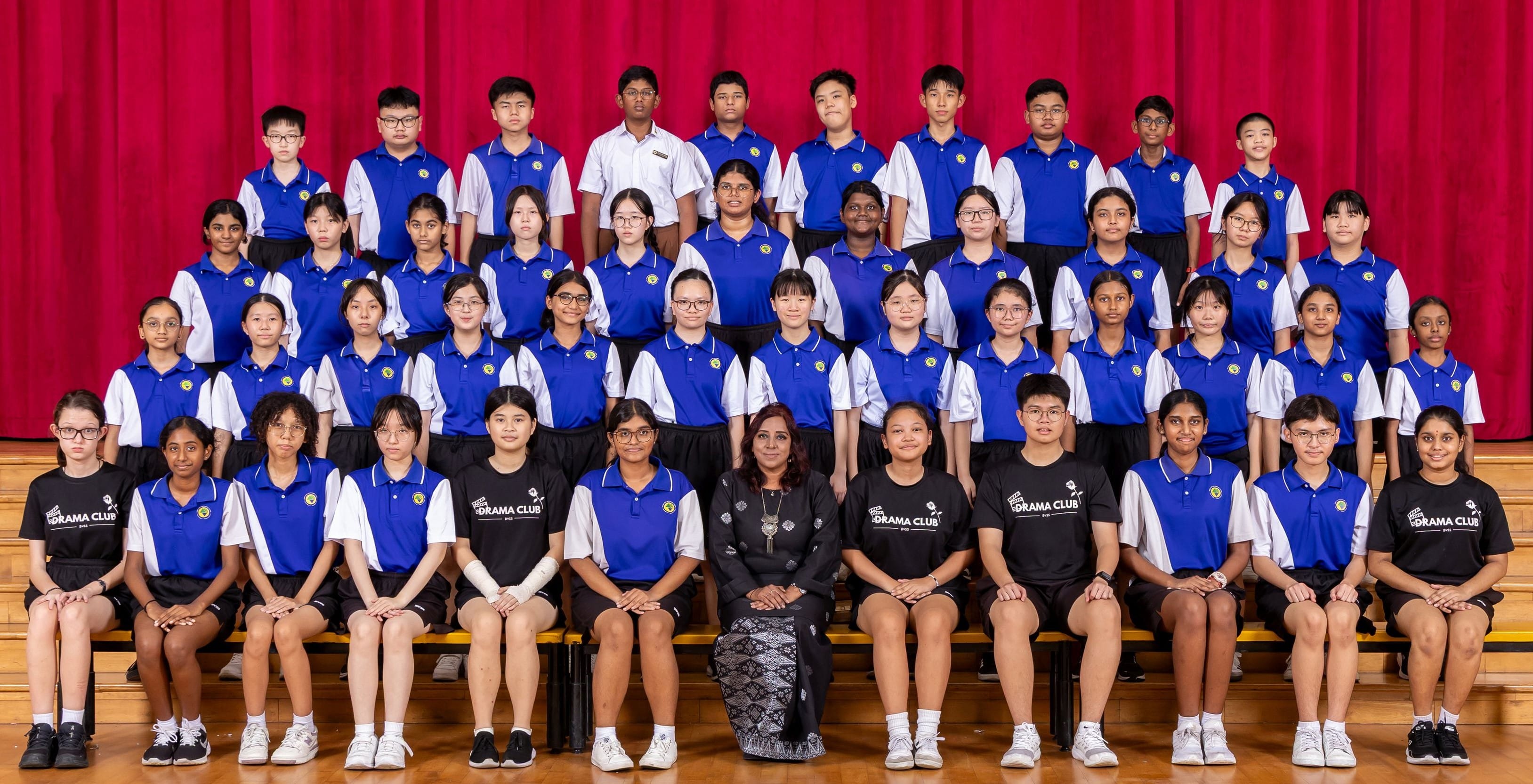 Large group of school students in blue uniforms and Drama Club shirts posing in rows with a teacher on a stage.