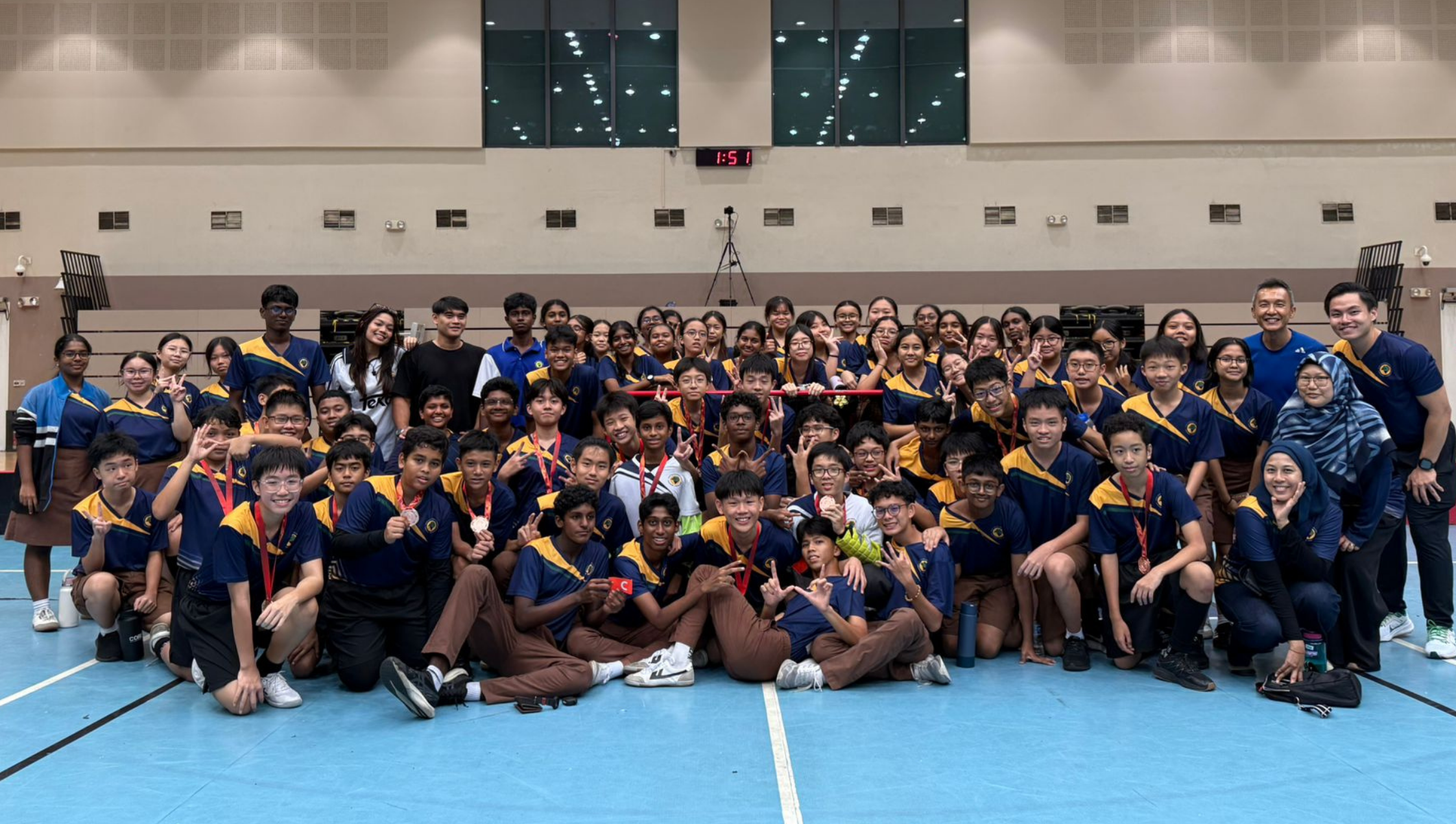Large group of students in matching blue uniforms pose with medals on a gym floor under a scoreboard.