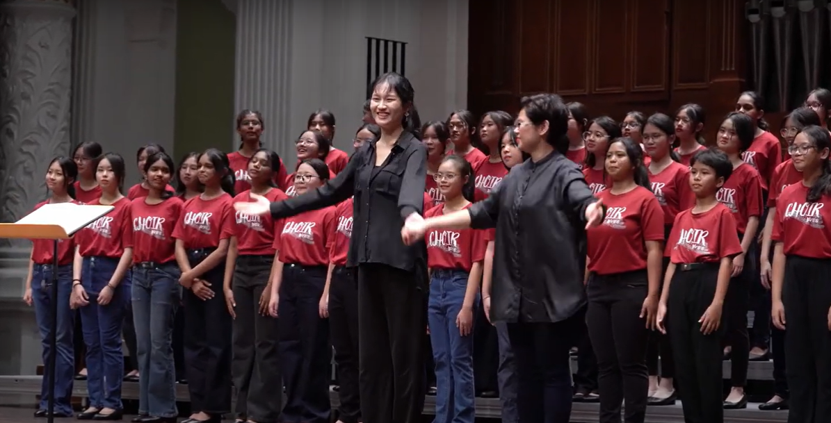 Two conductors in black lead a large youth choir wearing red shirts labeled "CHOIR" on a stage.
