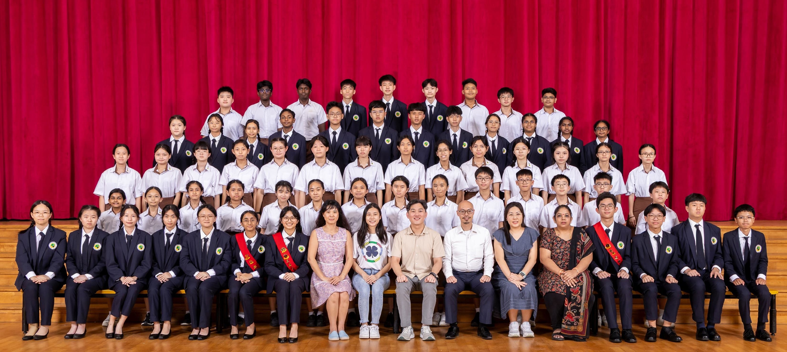 Large group of school students and teachers in uniforms posed in rows on a stage with a red curtain backdrop.