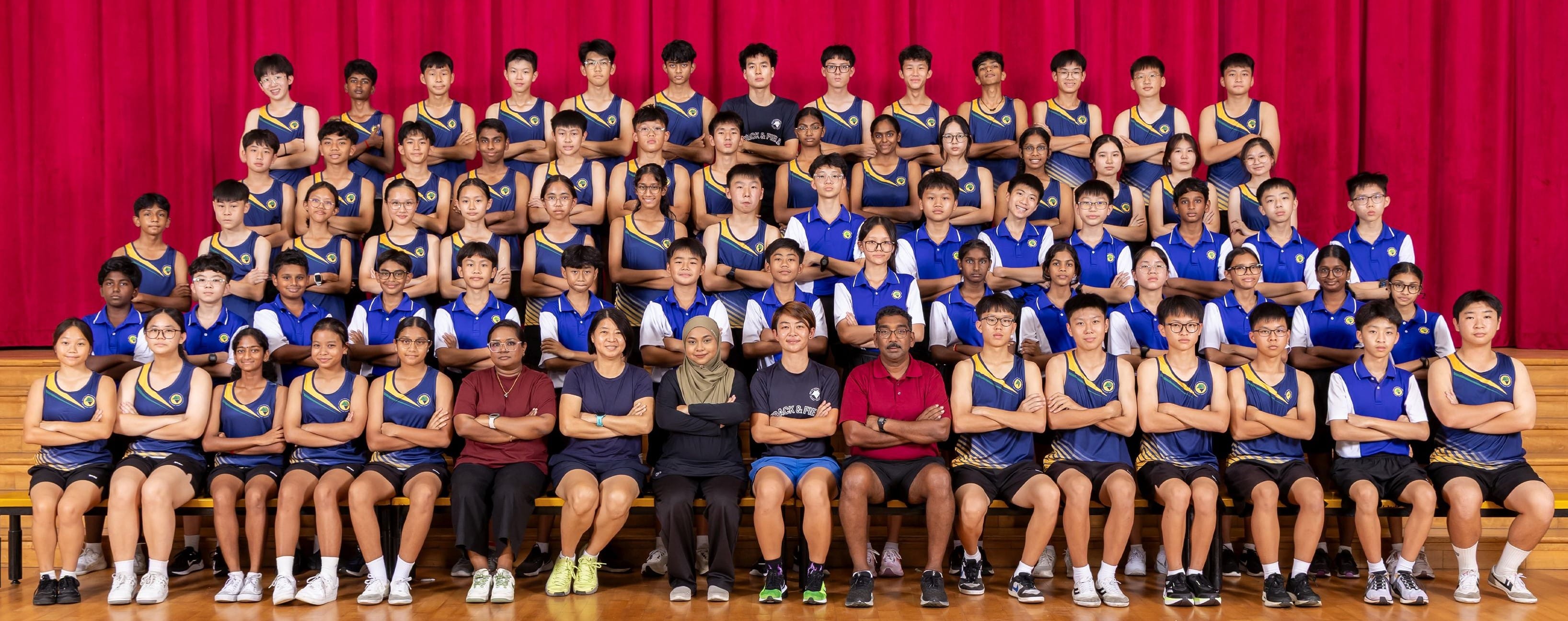 Large school track and field team posing in rows with arms crossed, wearing blue uniforms, in front of a red curtain.
