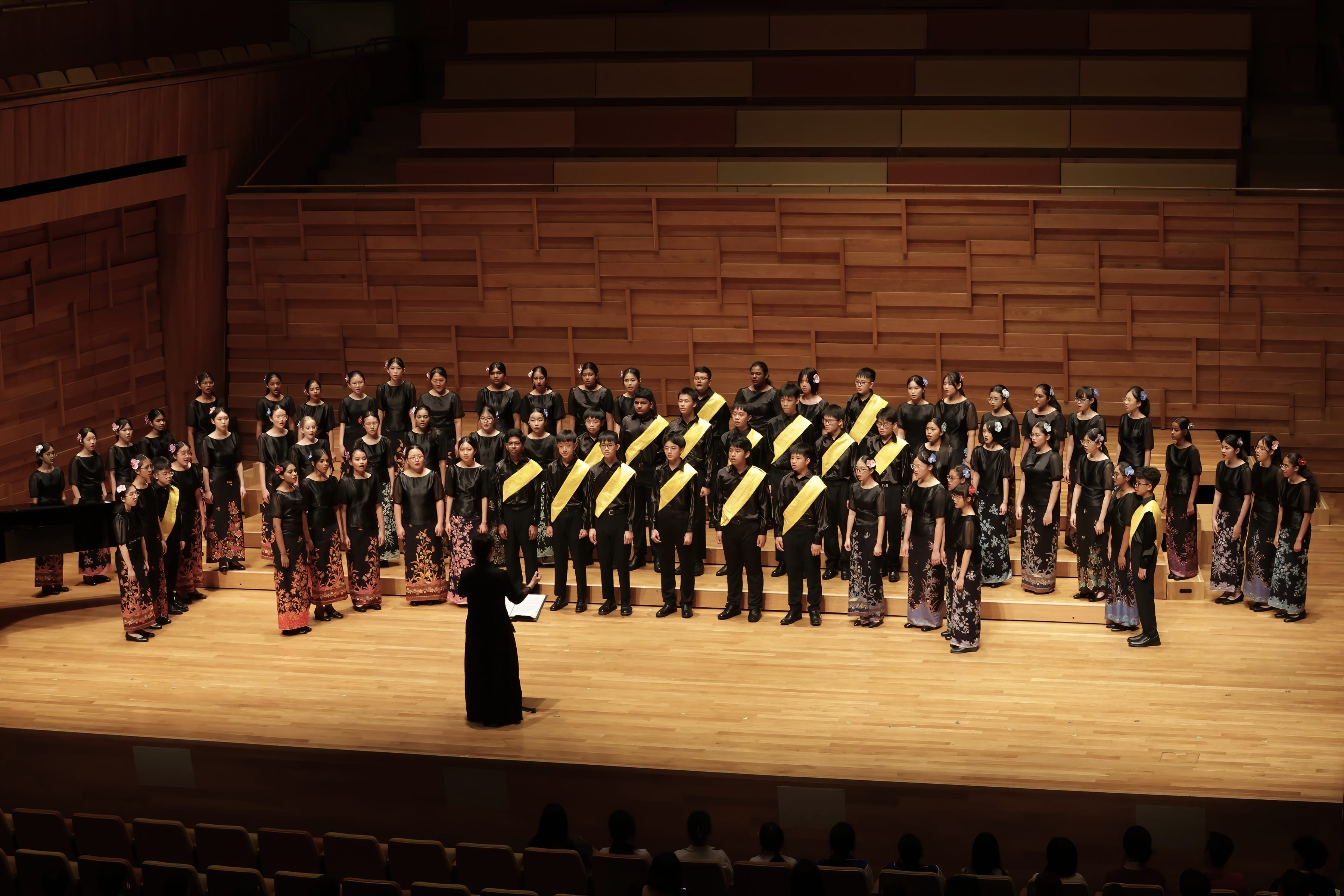 A large youth choir in black outfits performs on a wooden concert stage, led by a conductor facing them.