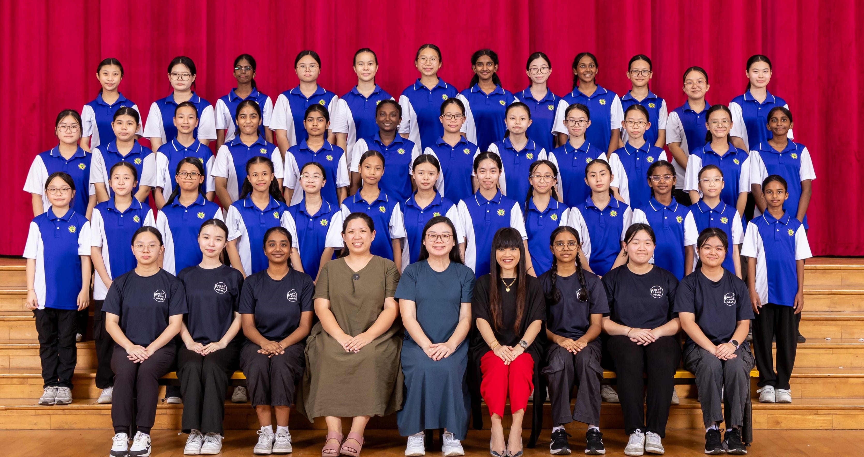 Large group of girls in blue and white uniforms posing with teachers on wooden bleachers before a red curtain.