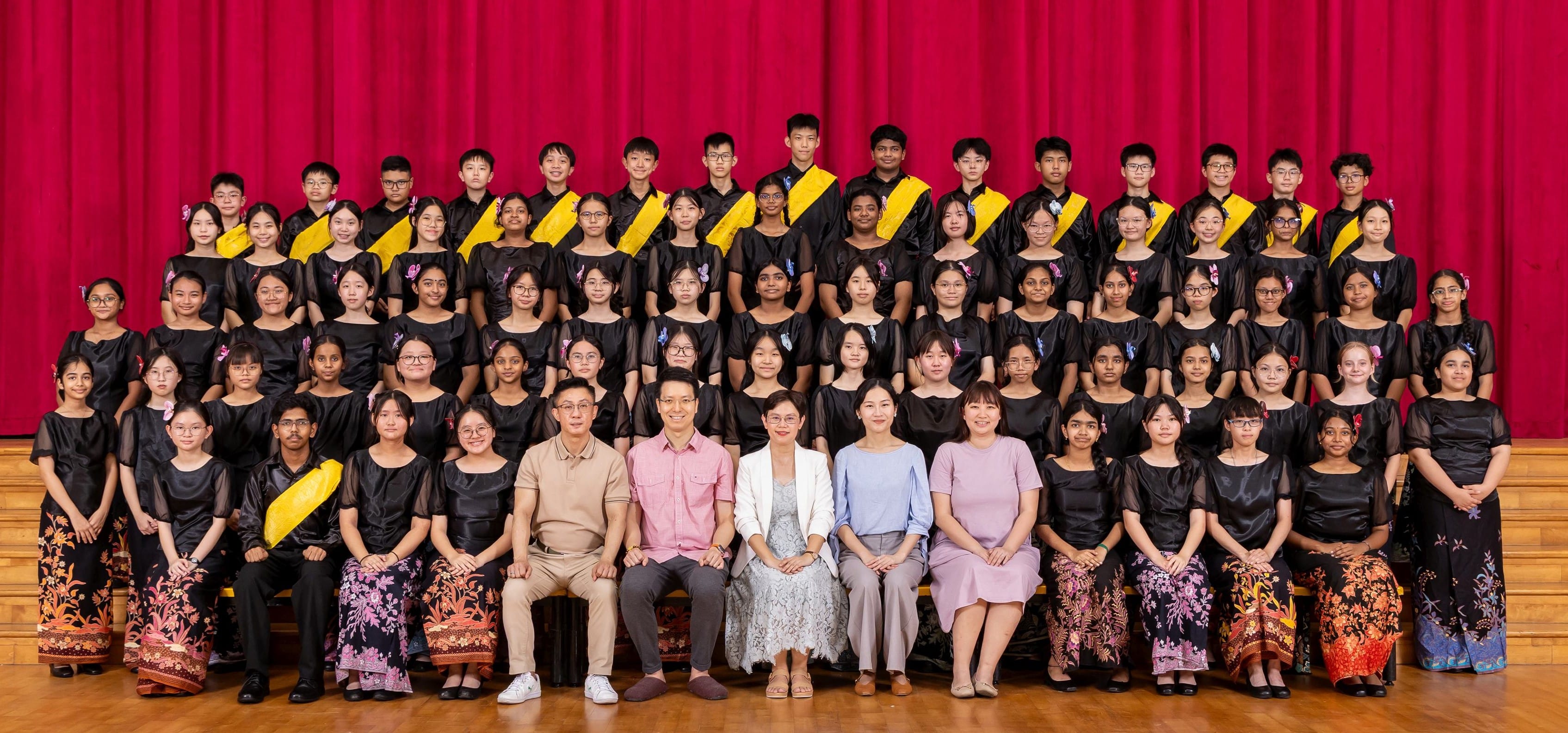 Large group of students in black uniforms with yellow sashes pose with five adults on a stage with a red curtain.