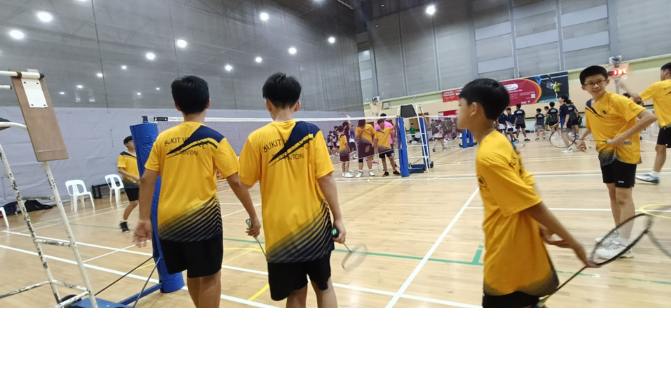 Boys in yellow Bukit badminton jerseys holding rackets stand on an indoor court during a tournament.