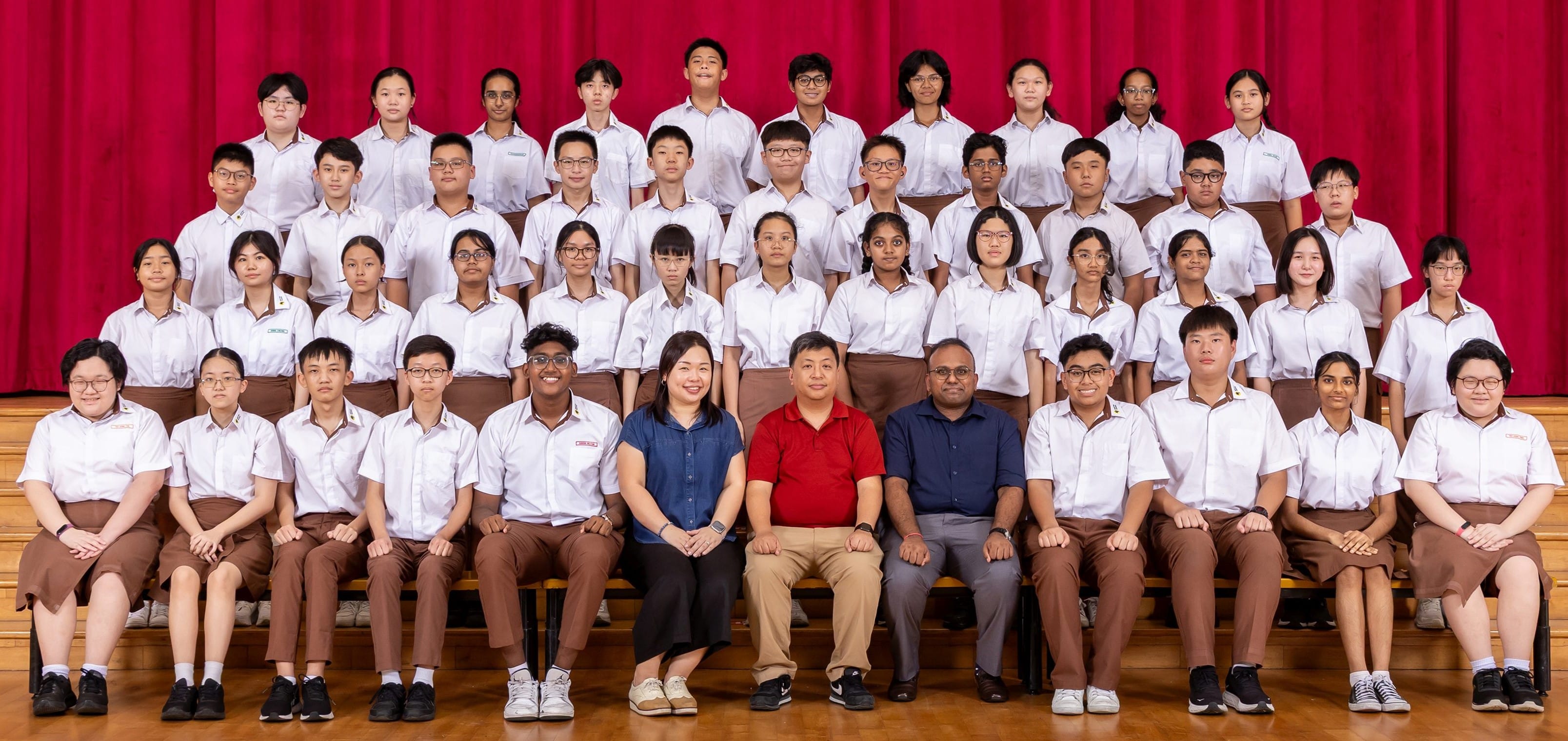 Large school group photo with students in white uniforms and three teachers seated in front, posing before a red curtain.