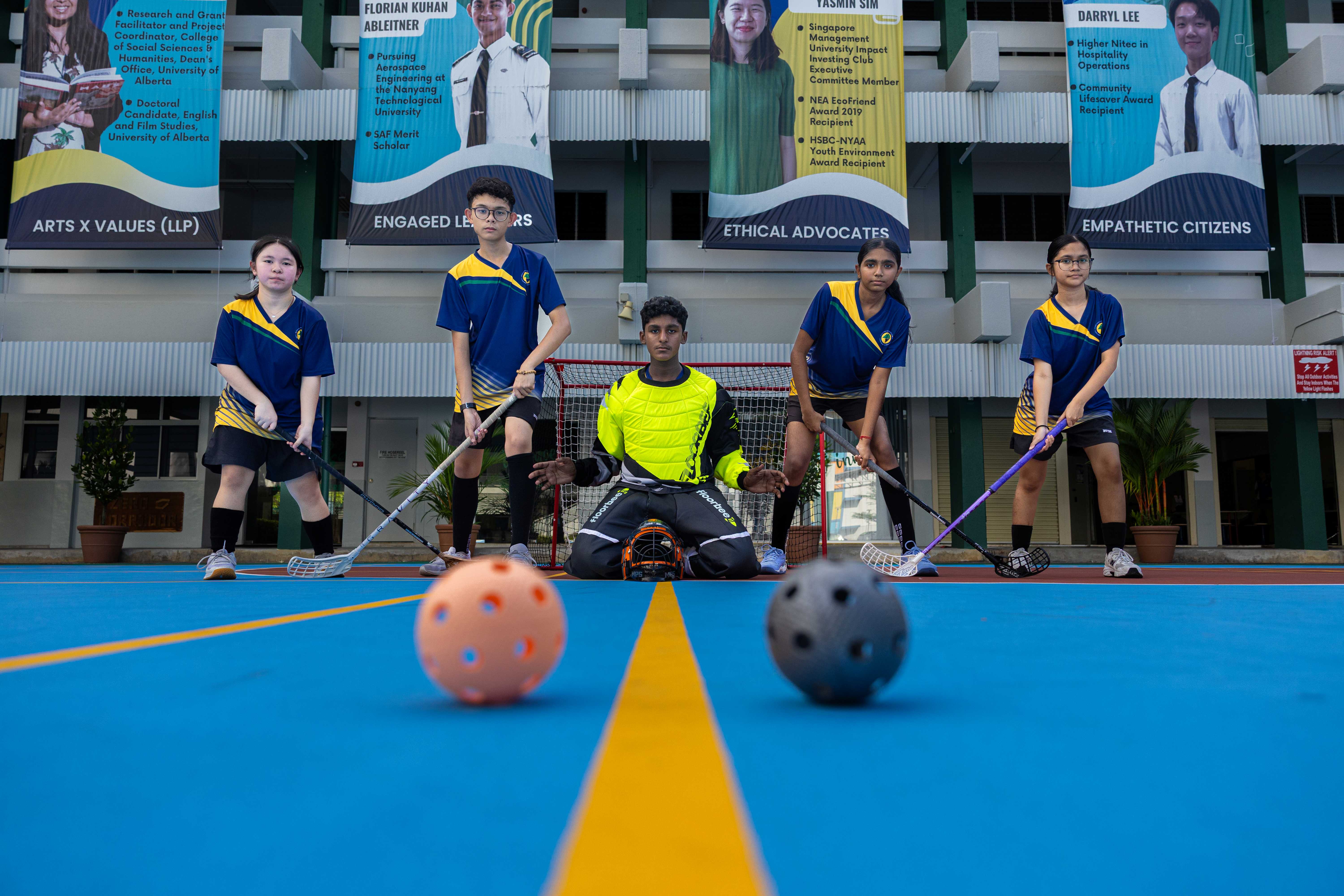 Five students in blue jerseys pose on a floorball court, with a goalkeeper kneeling at a small goal and two balls in foreground.
