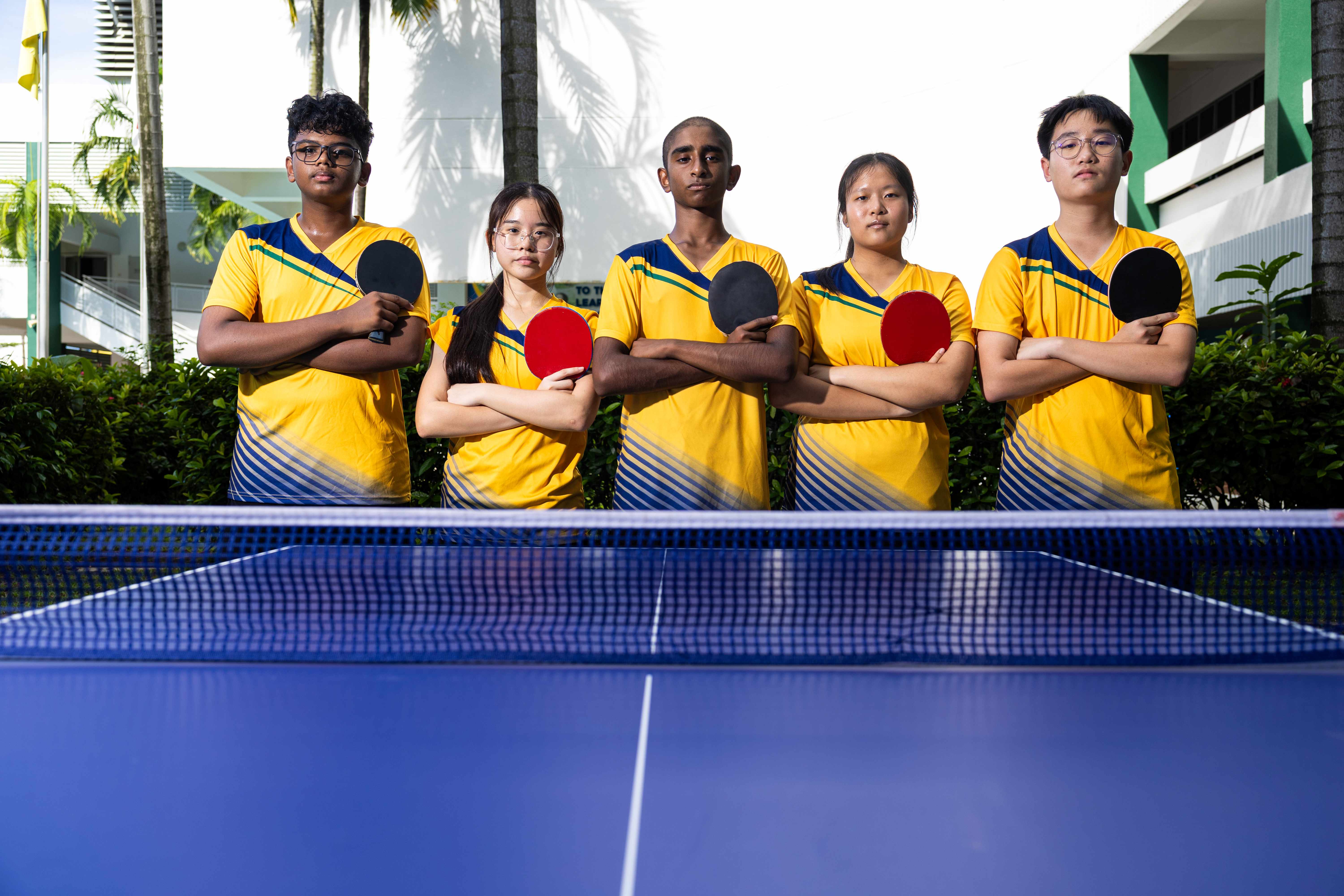 Five youth in matching yellow jerseys hold table tennis paddles with arms crossed behind a blue ping pong table outdoors.
