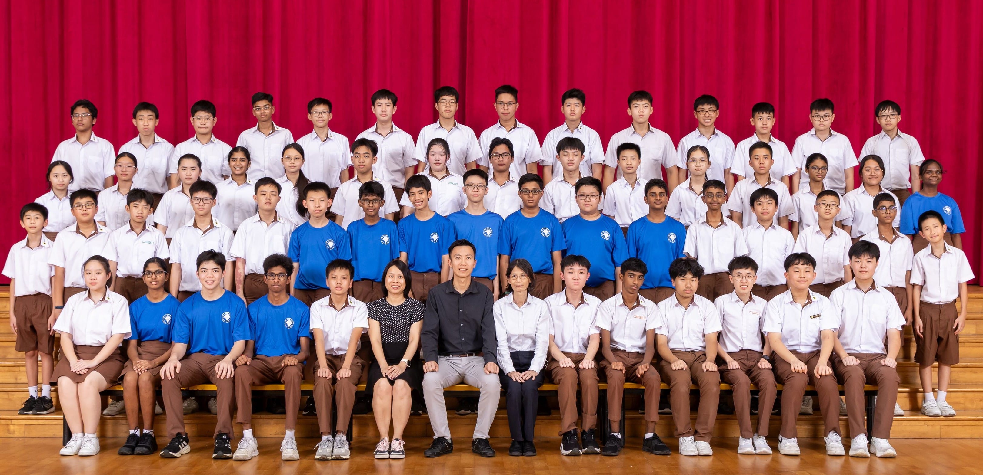 Large group of students in white and blue uniforms posing with two adults in front of a red curtain.