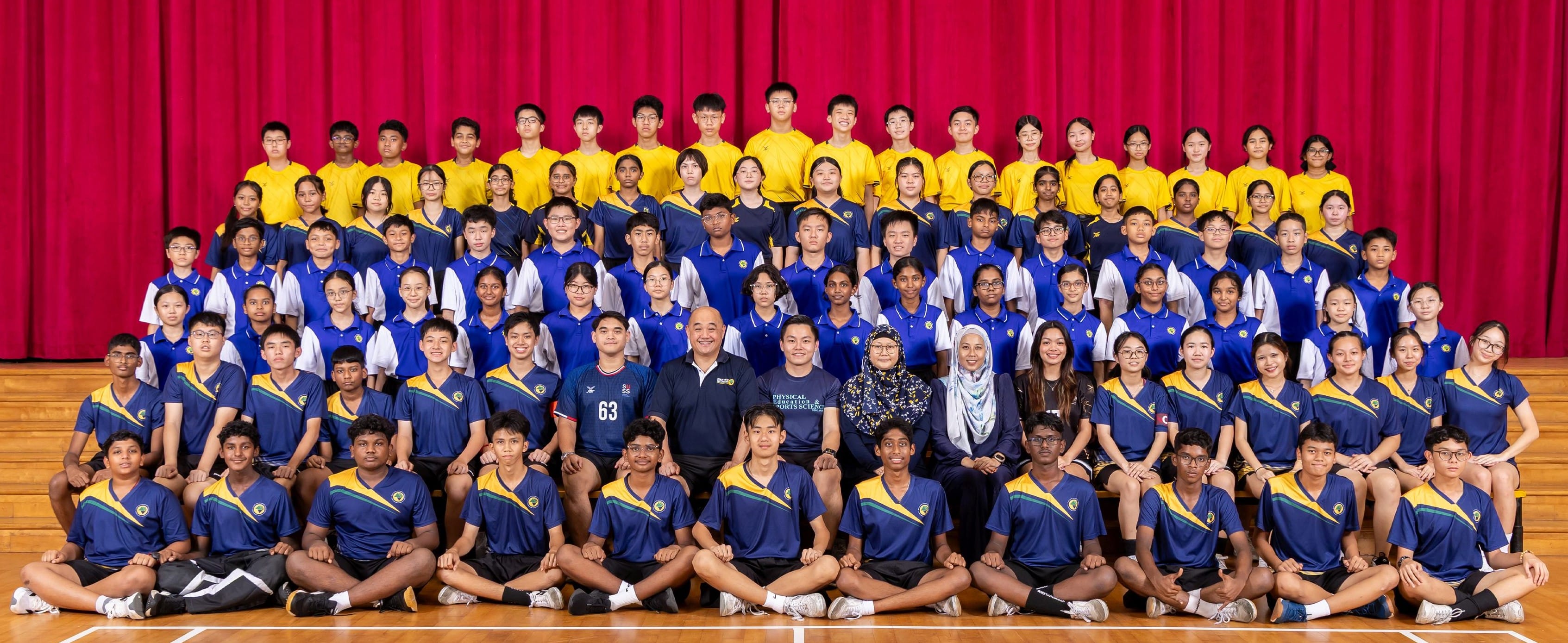 Large group of students and teachers in blue and yellow uniforms posing for a group photo in a school hall.