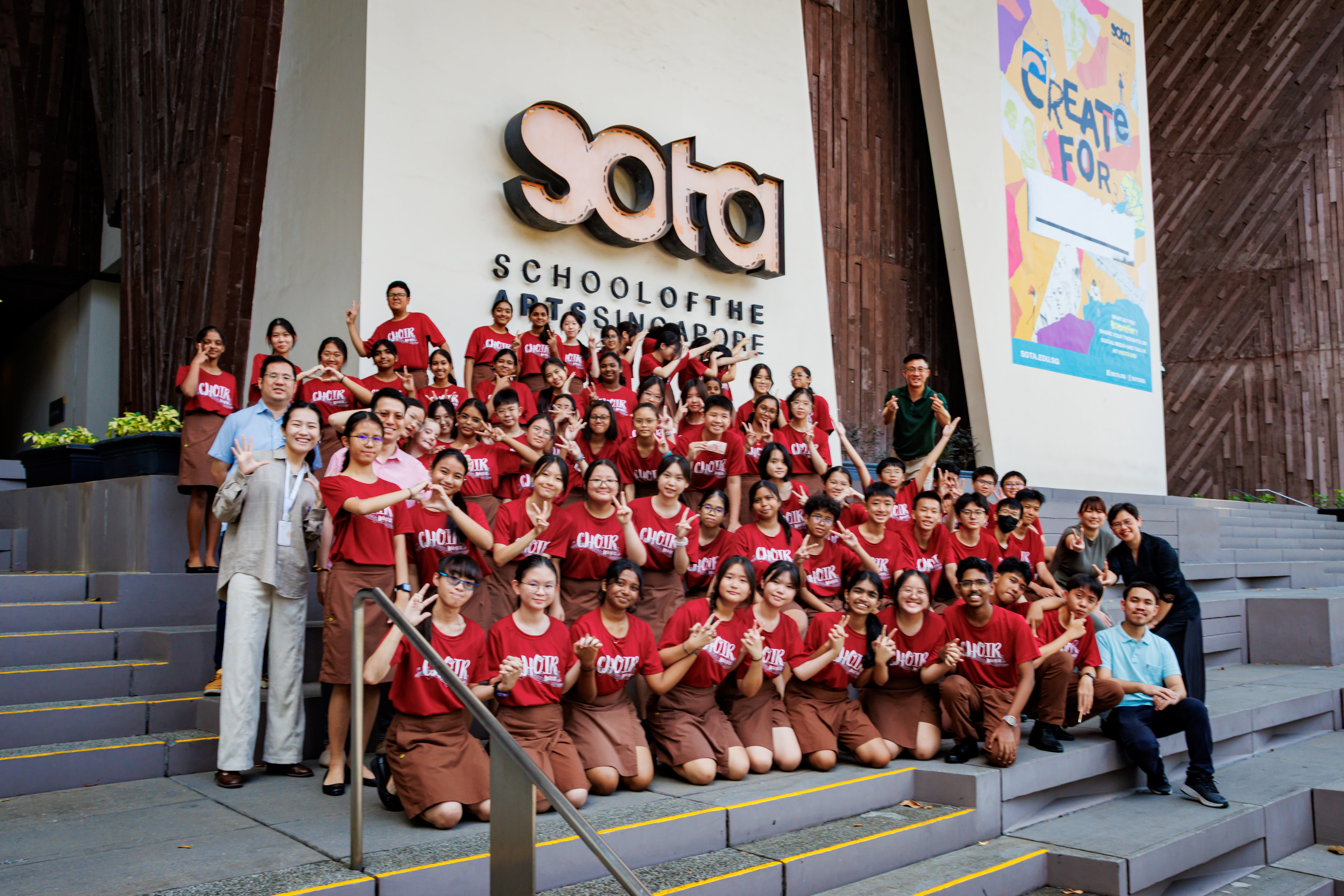 Large group in red choir shirts posing on steps in front of a building sign reading Sota.
