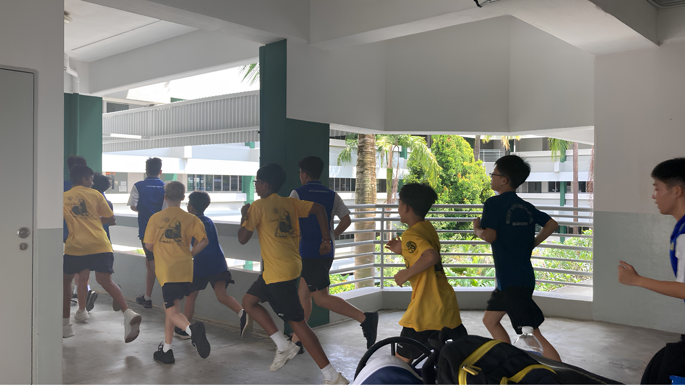 A group of boys in yellow and blue shirts running along an outdoor school corridor with green trees visible outside.