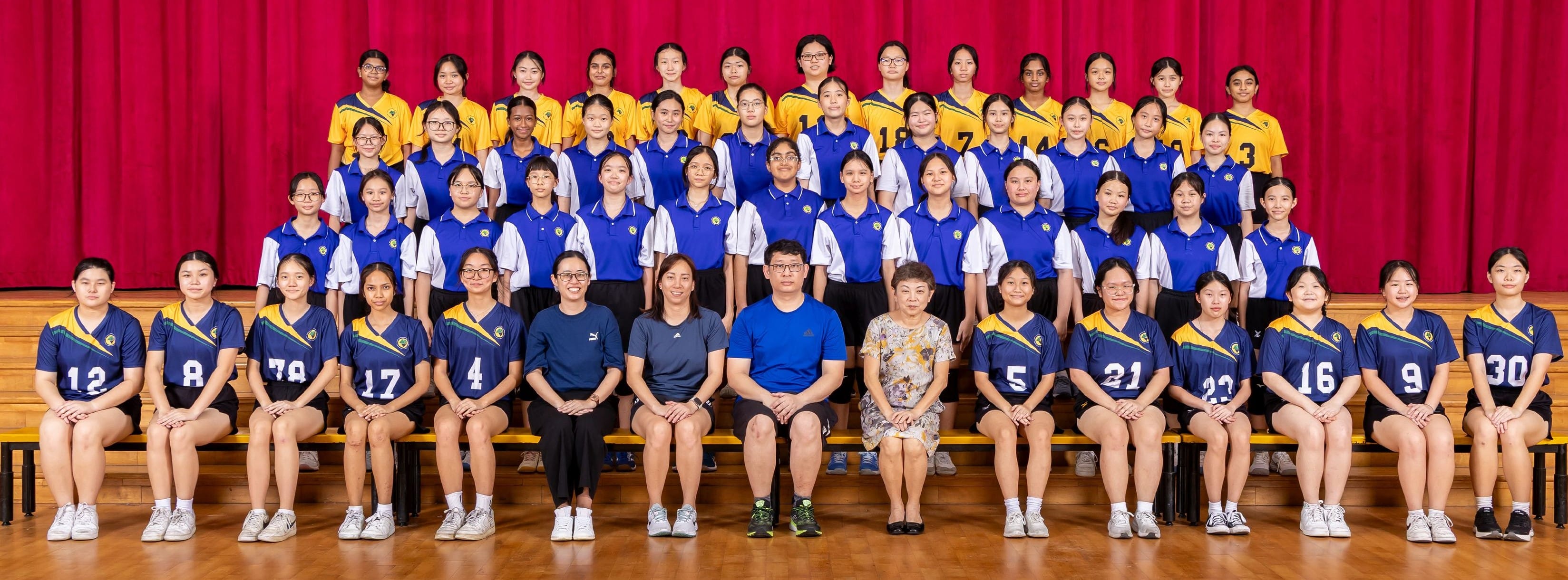 Large group of female students in blue and yellow sports uniforms posed in rows with coaches before a red curtain.