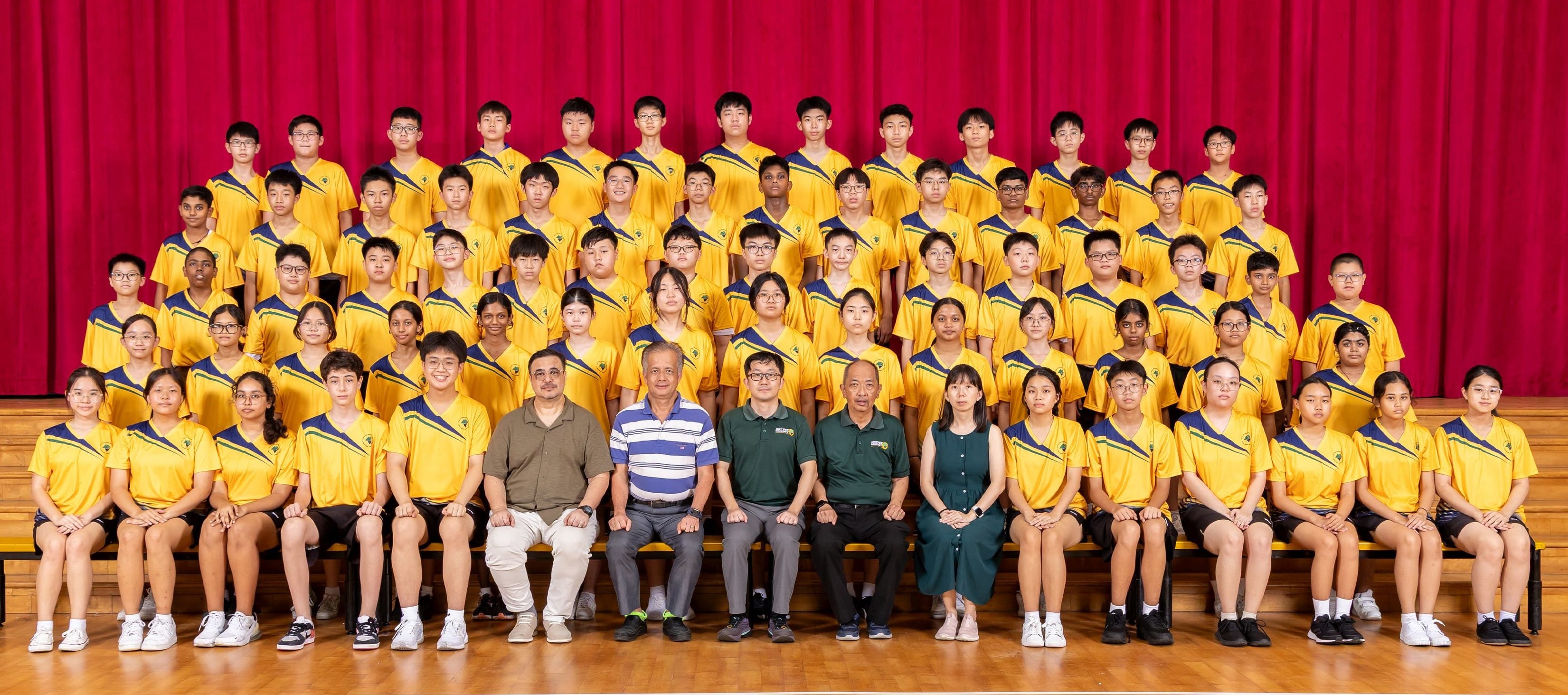 Large group of students in yellow uniforms posing with several adults in front of a red curtain backdrop.